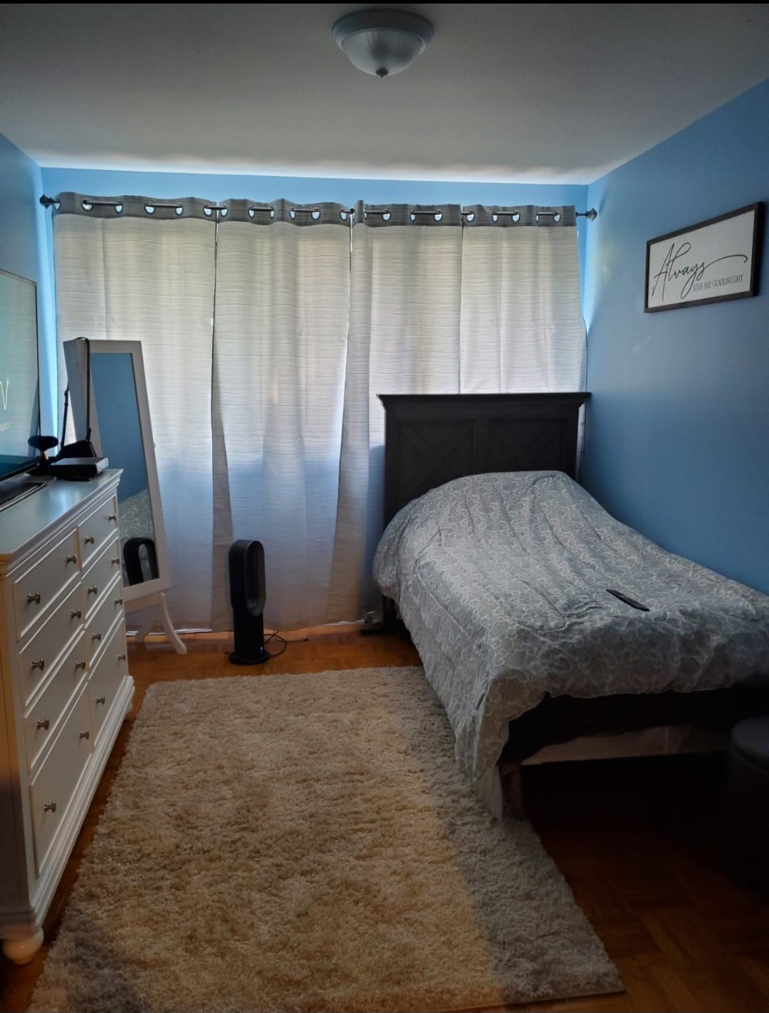 A bedroom with light blue walls, a bed with a patterned bedspread, a white dresser, a mirror, a TV on a stand, a fan, a black remote on the bed, a beige rug, curtains covering a window, and a framed wall art that reads "Always kiss me goodnight."