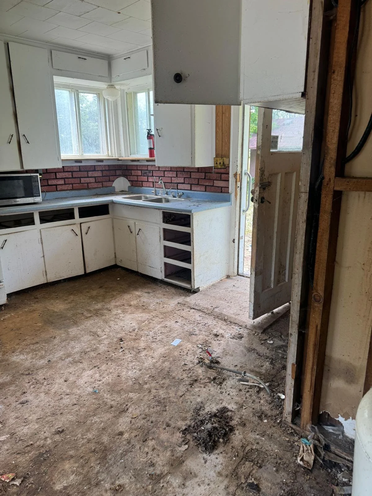 Kitchen under renovation with missing floor, dirty cabinets, and a door leading outside.