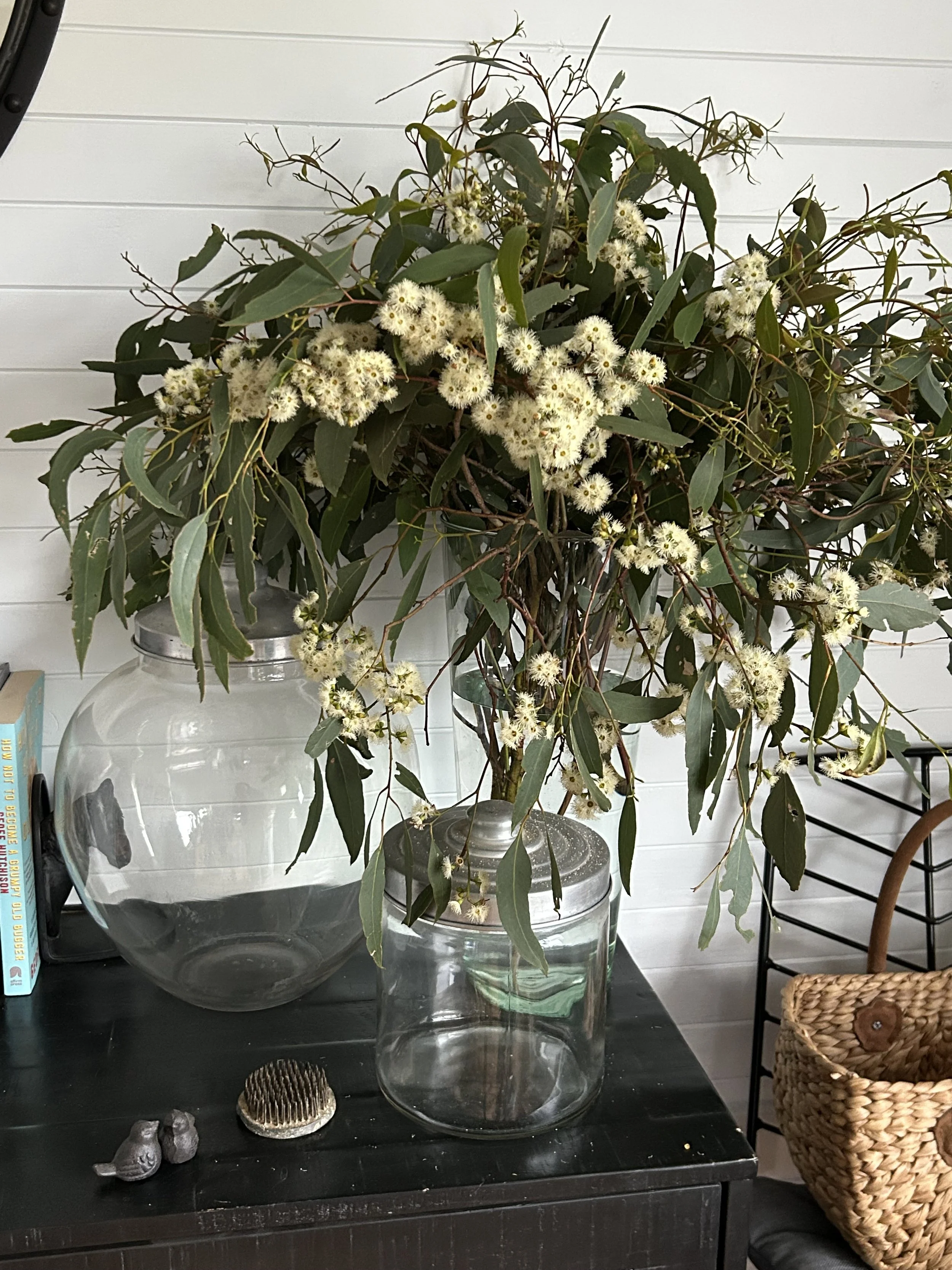 A floral arrangement with White fluffy flowers and green leaves in a glass vase on a black table, with decorative items including small bird figurines, a hairbrush, and a woven basket nearby.