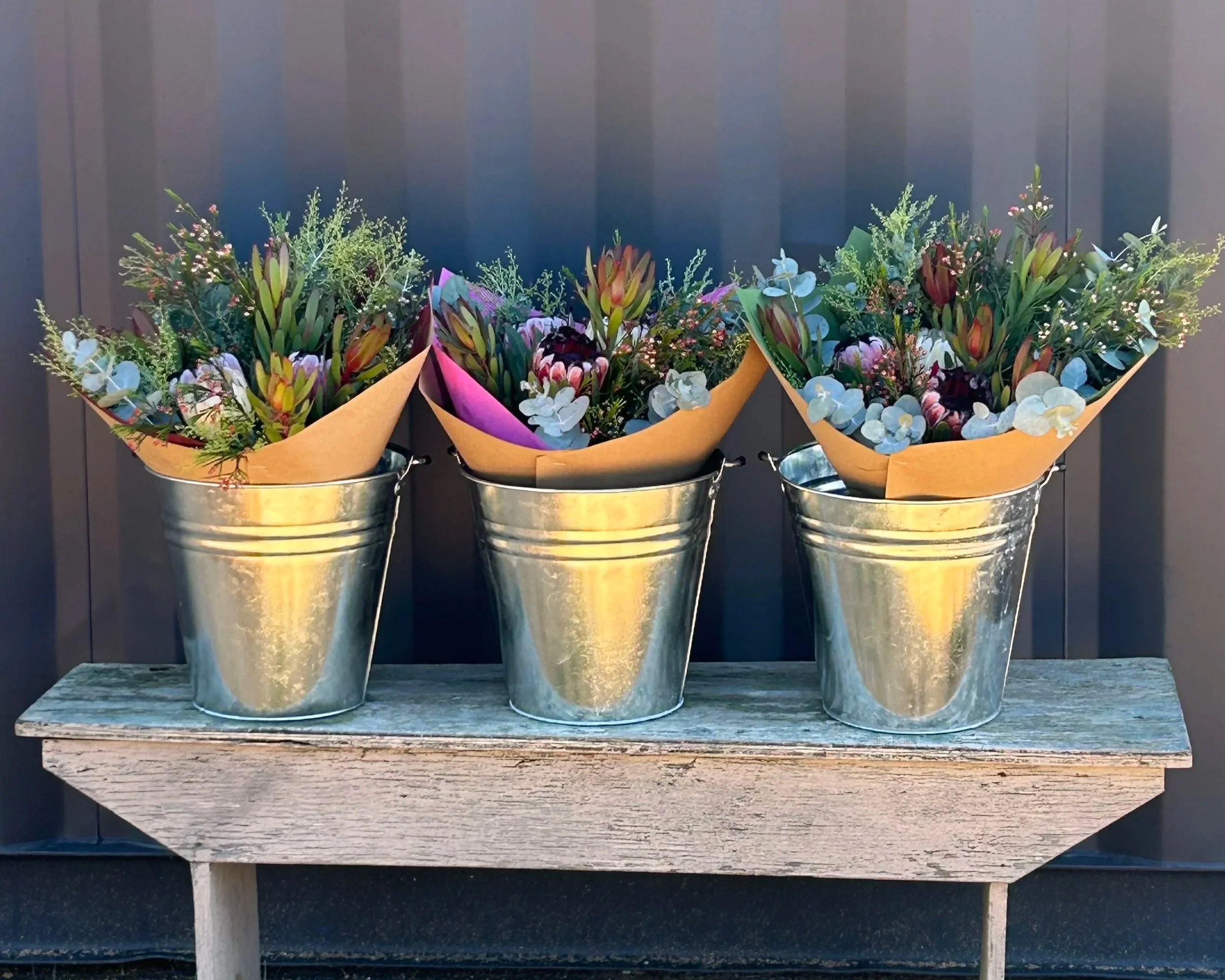 Four metal buckets filled with assorted flowers placed on a weathered wooden table against a dark background.