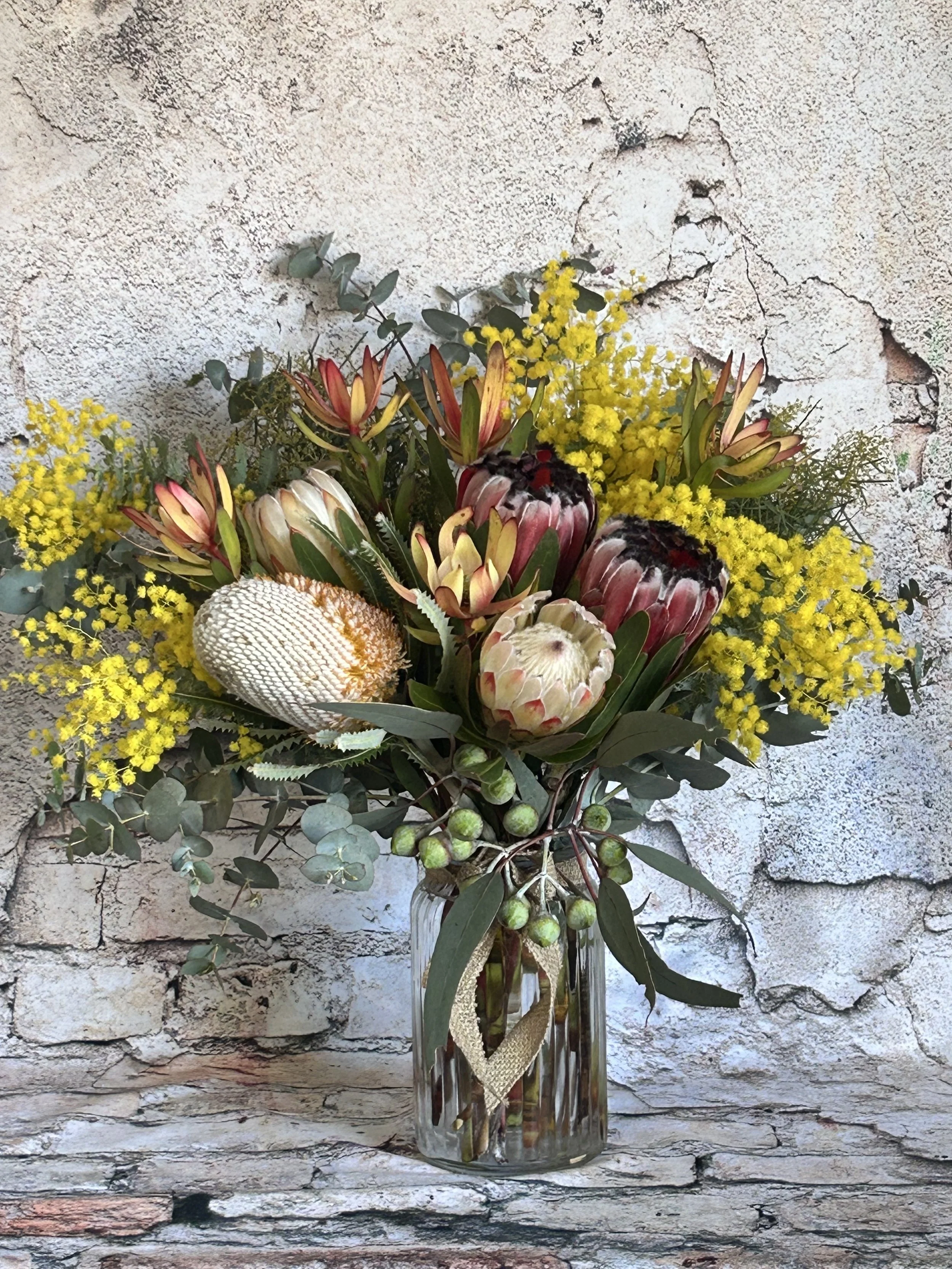 A floral arrangement with various flowers and greenery in a tall glass vase, set against a textured stone wall background.