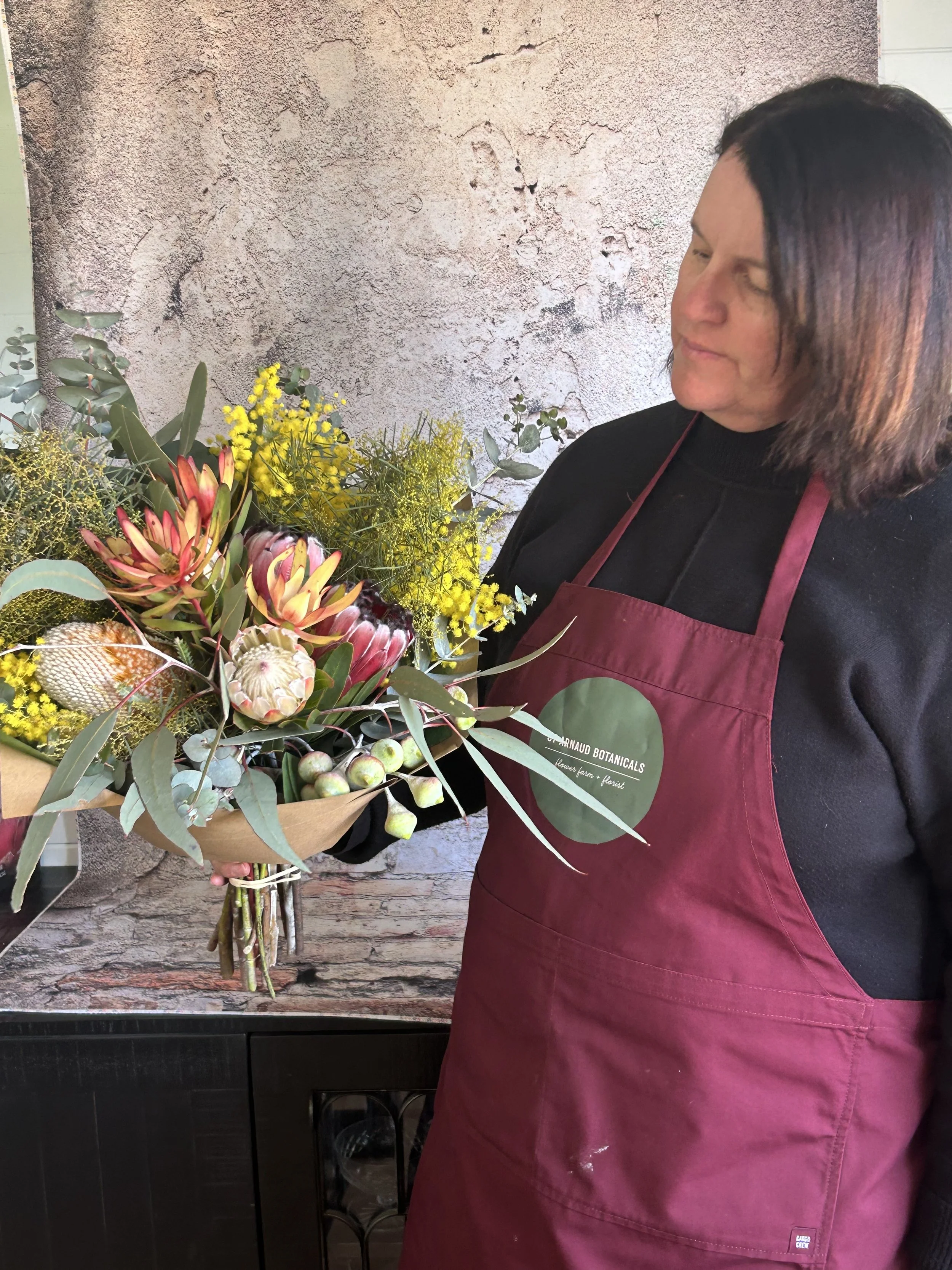 A woman standing with her eyes closed, wearing a black shirt and a burgundy apron with the logo of 'Gaurnaud Botanicals,' holding a bouquet of various colorful flowers and greenery against a textured wall background.