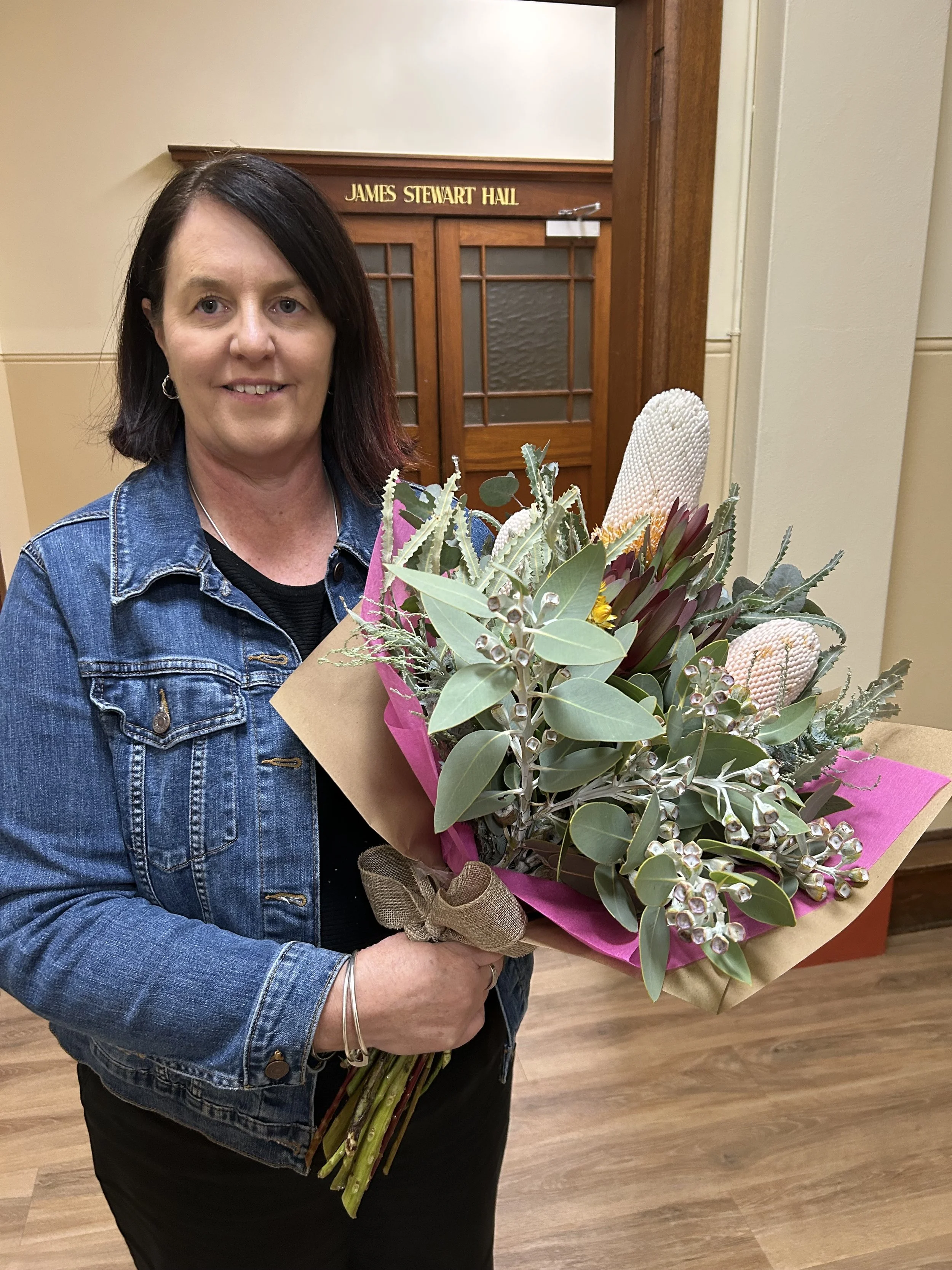 A woman holding a bouquet of flowers in front of a wooden door labeled 'James Stewart Hall'.