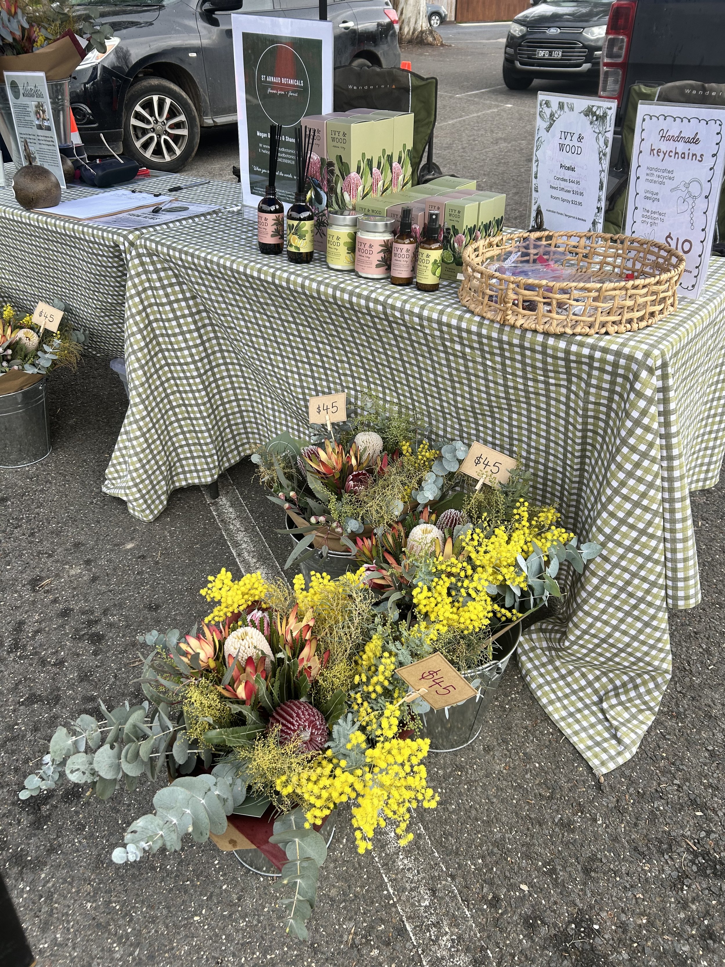 Outdoor market stall with floral arrangements, plants, and handmade products displayed on a checkered tablecloth, with parking lot and cars in the background.