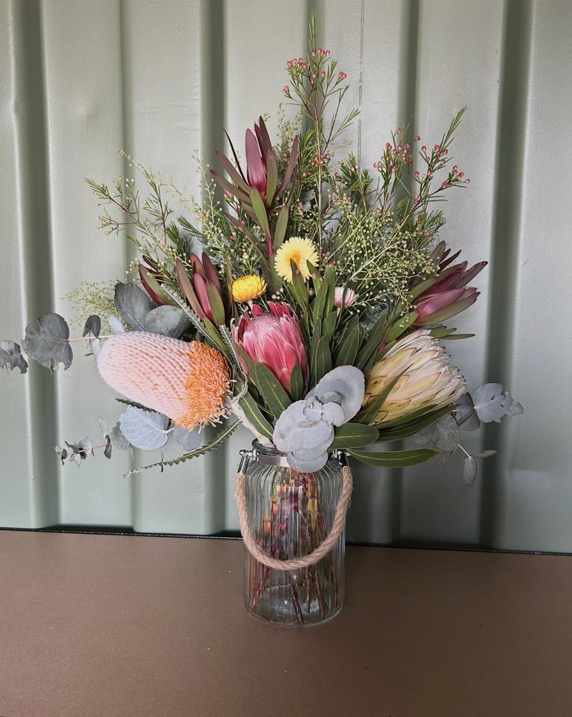 A colorful flower bouquet in a clear glass vase with a rope handle, placed on a brown surface against a green wall background.