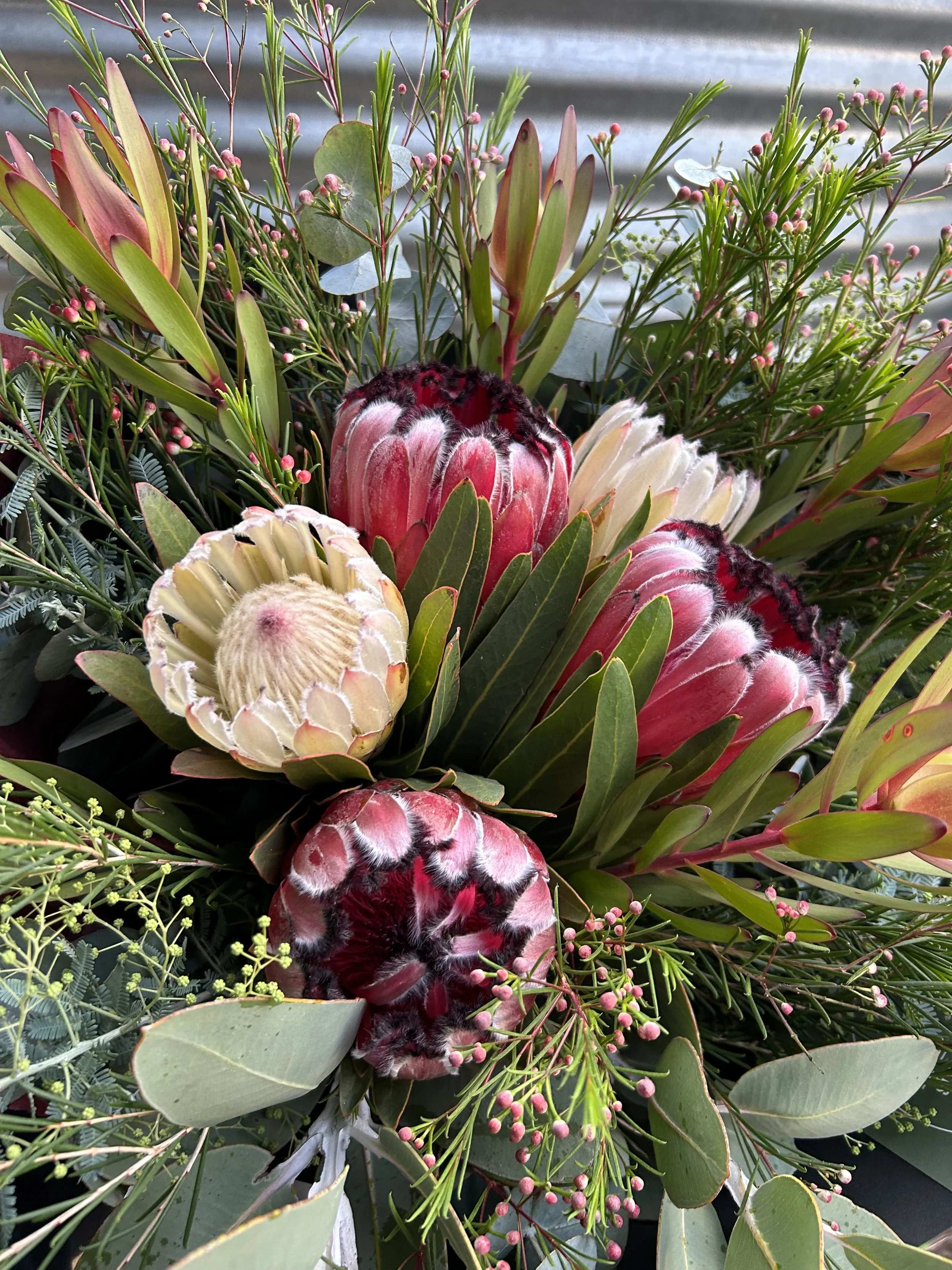 A bouquet of various flowers and greenery, including Protea flowers, surrounded by green leaves and small pink buds.