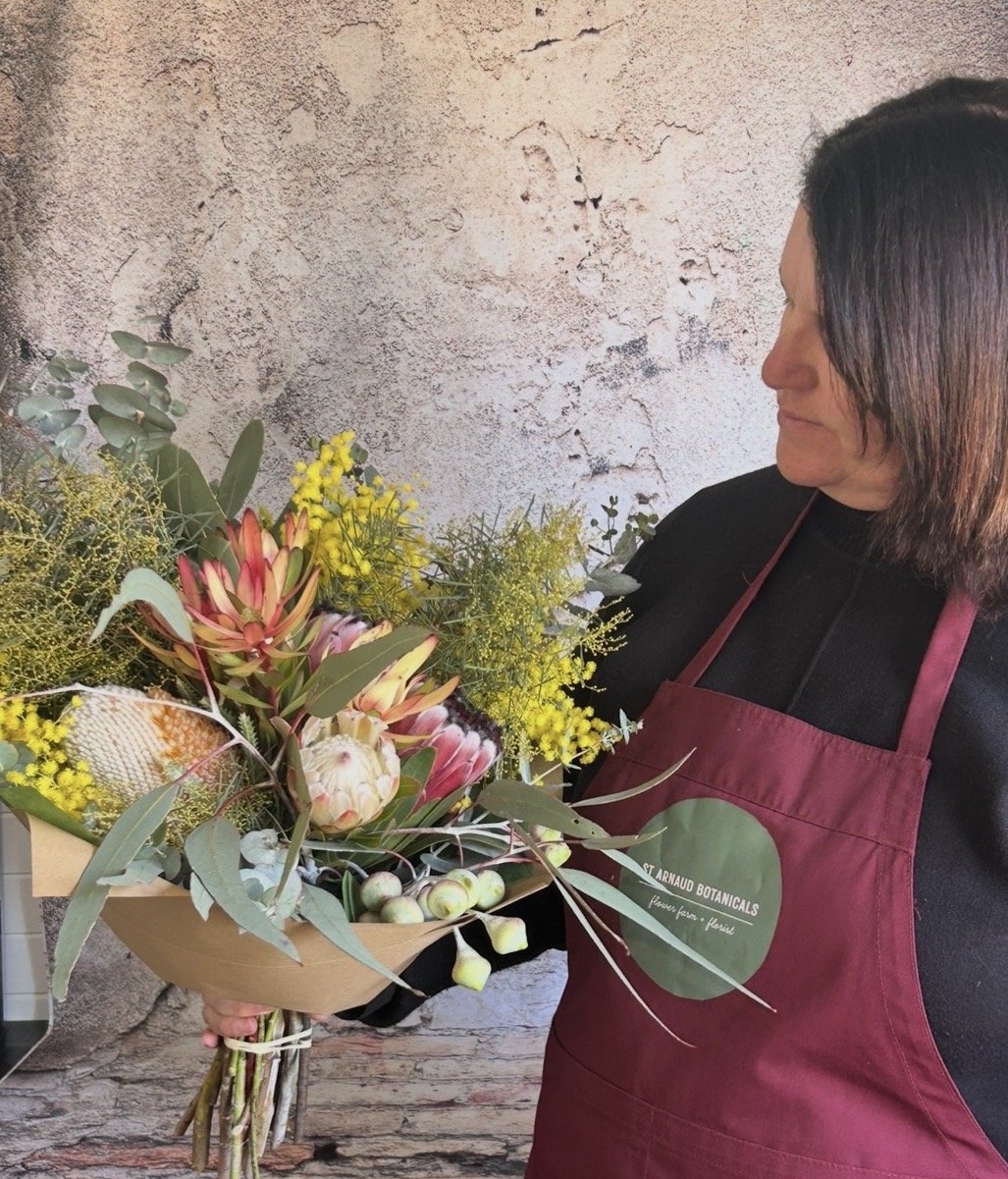 A woman wearing a burgundy apron holding a large bouquet of various flowers and greenery, standing in front of a textured wall.