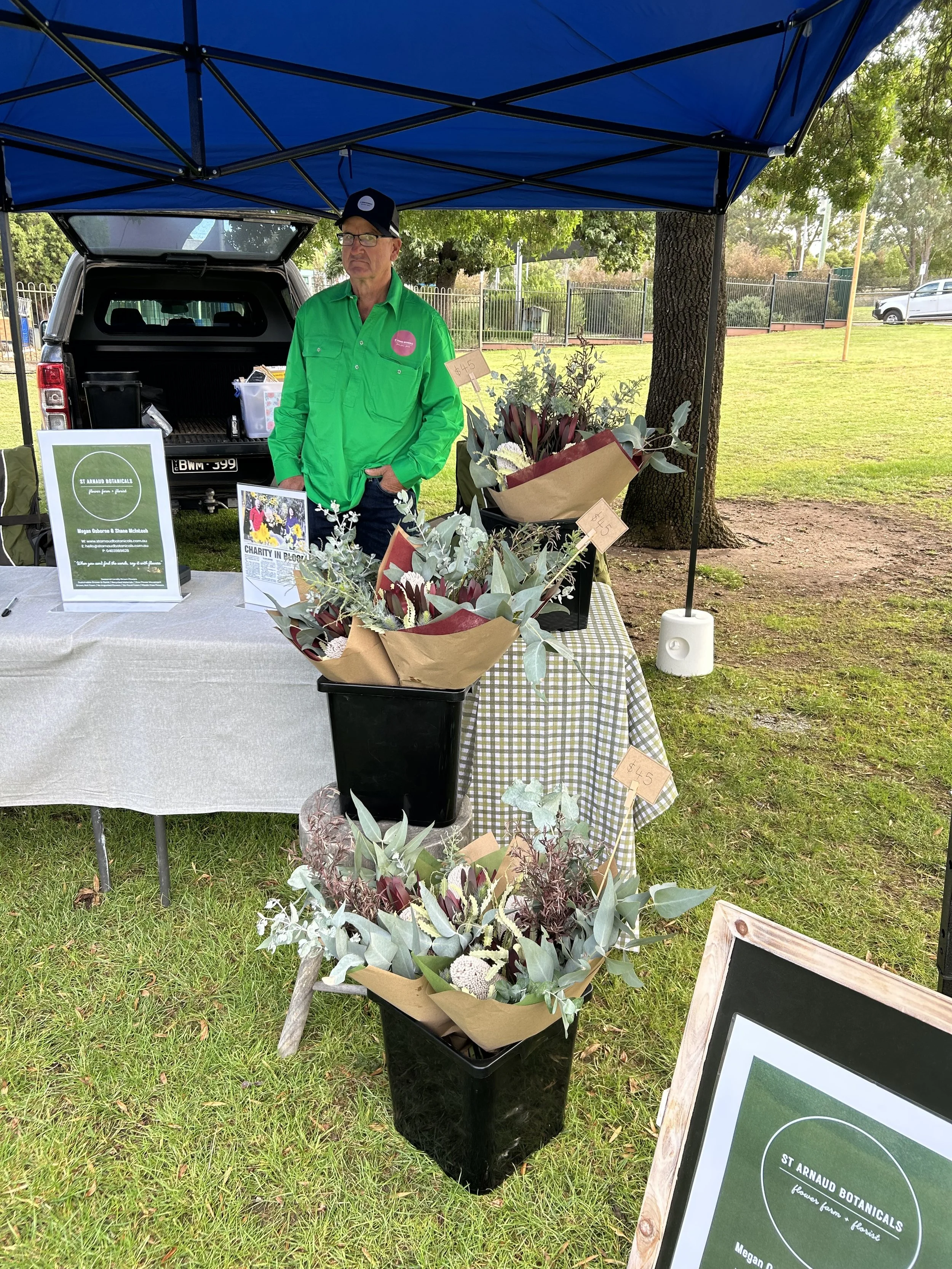 Man standing under a blue canopy at a plant stand in a park, with flower bouquets on display and a sign with the text 'ST ARNAUD BOTANICALS.'