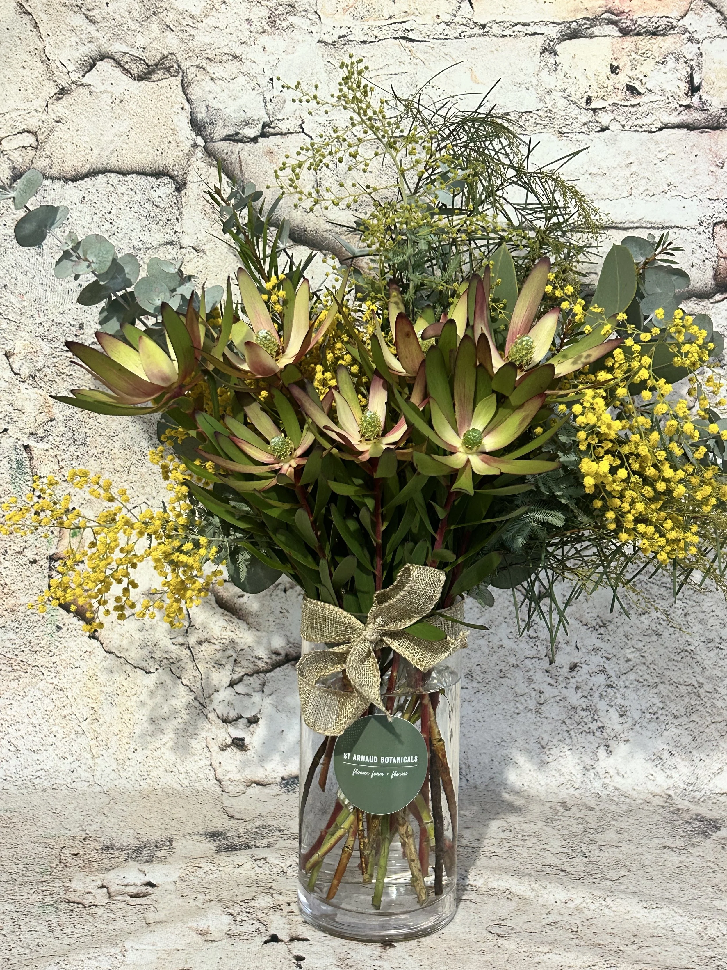 Bouquet of yellow and green flowers in a glass vase, tied with a gold ribbon, against a white textured wall background.