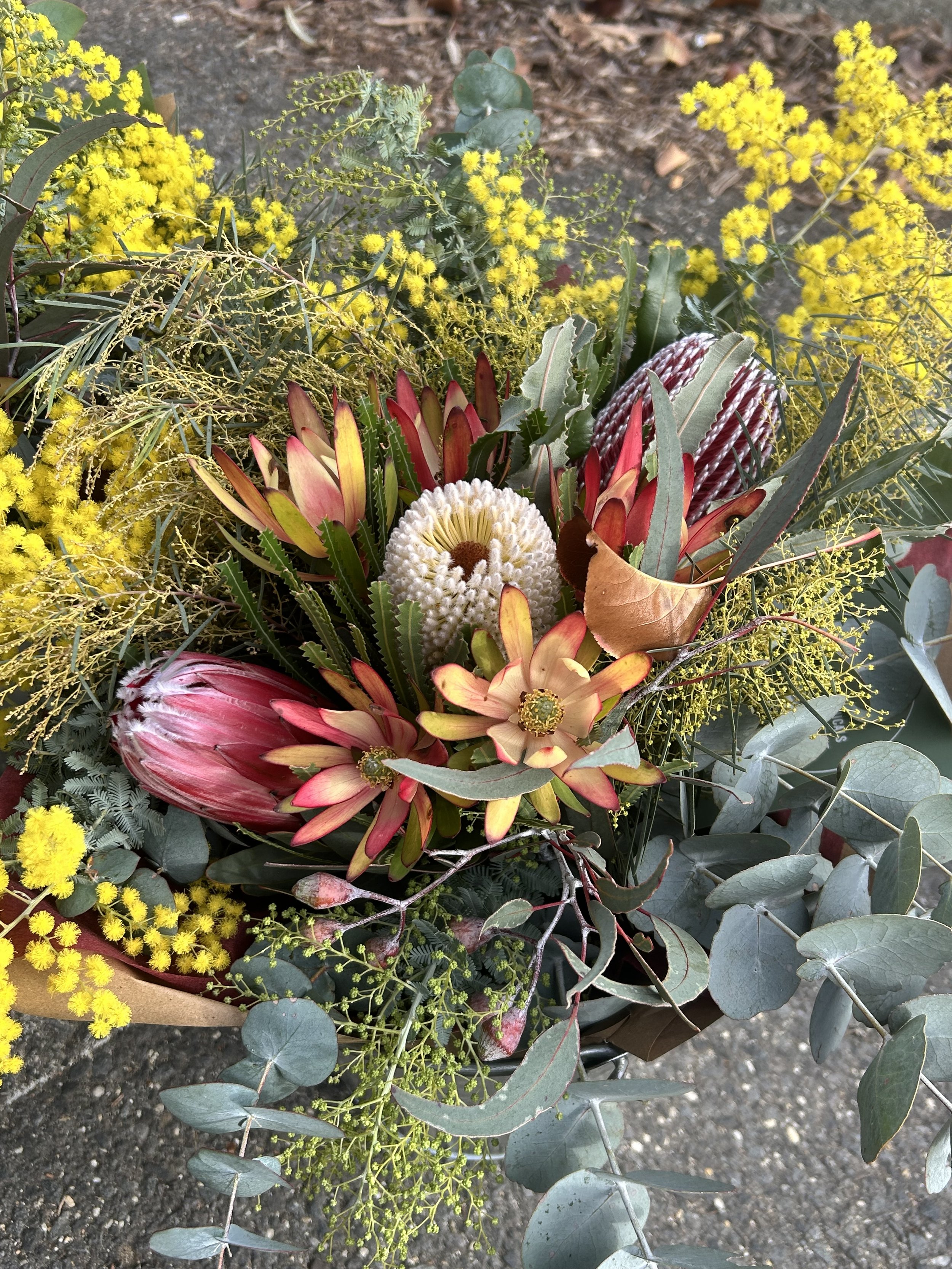 A bouquet of various flowers including yellow, pink, red, white, and green foliage, placed on a dark gravel surface.