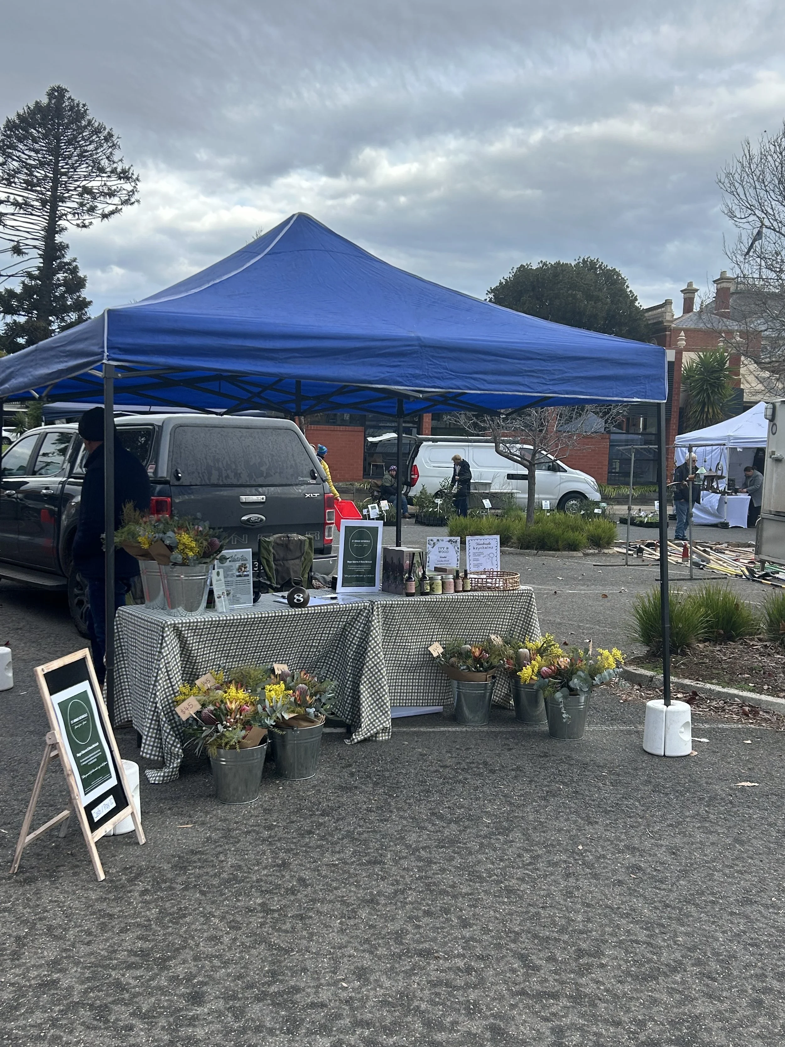An outdoor market scene with booths under tents, one blue and one white, selling flowers and plants. Several people are browsing and browsing, with cars parked in the background and a large brick building with chimneys visible. The sky is cloudy.