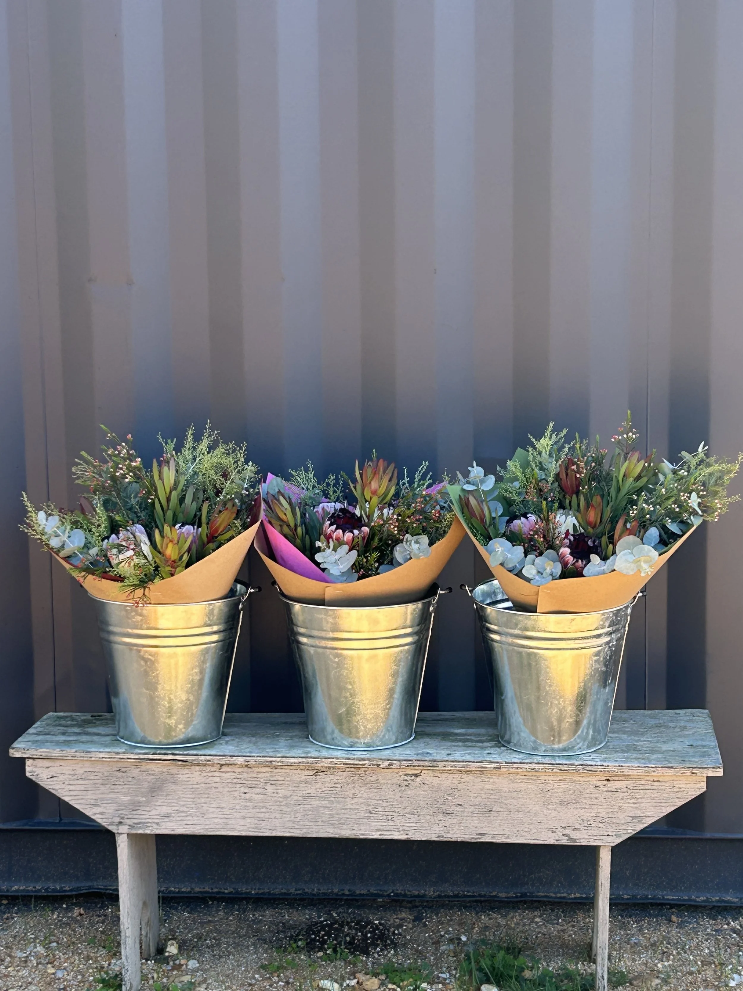 Three metal buckets filled with colorful flower bouquets placed on a wooden table in front of a dark wall with vertical paneling.