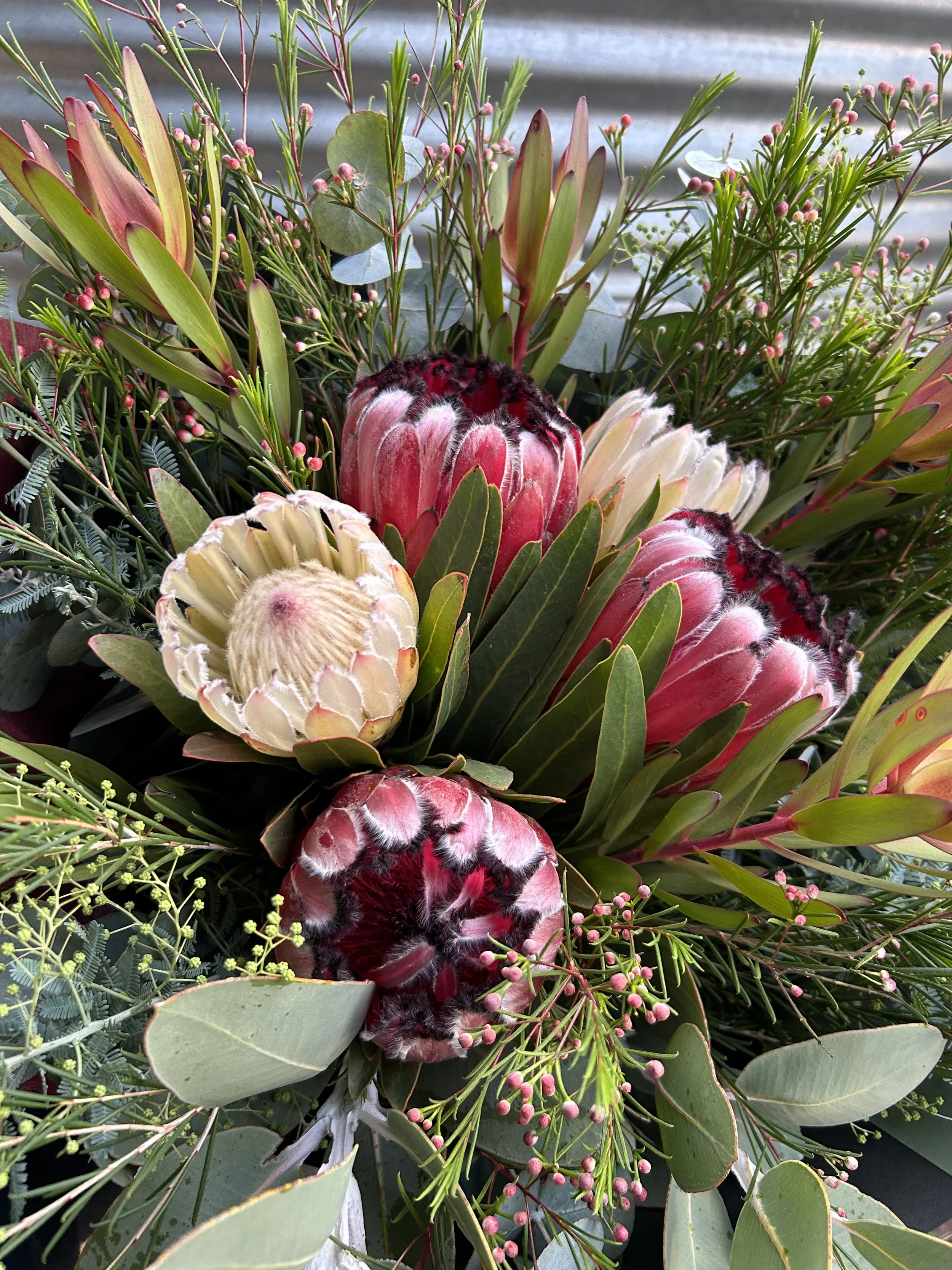 A lush bouquet of various green plants and flowers, including some pink-tipped Protea flowers, small pink buds, and green leaves with some grayish-green foliage in the background.