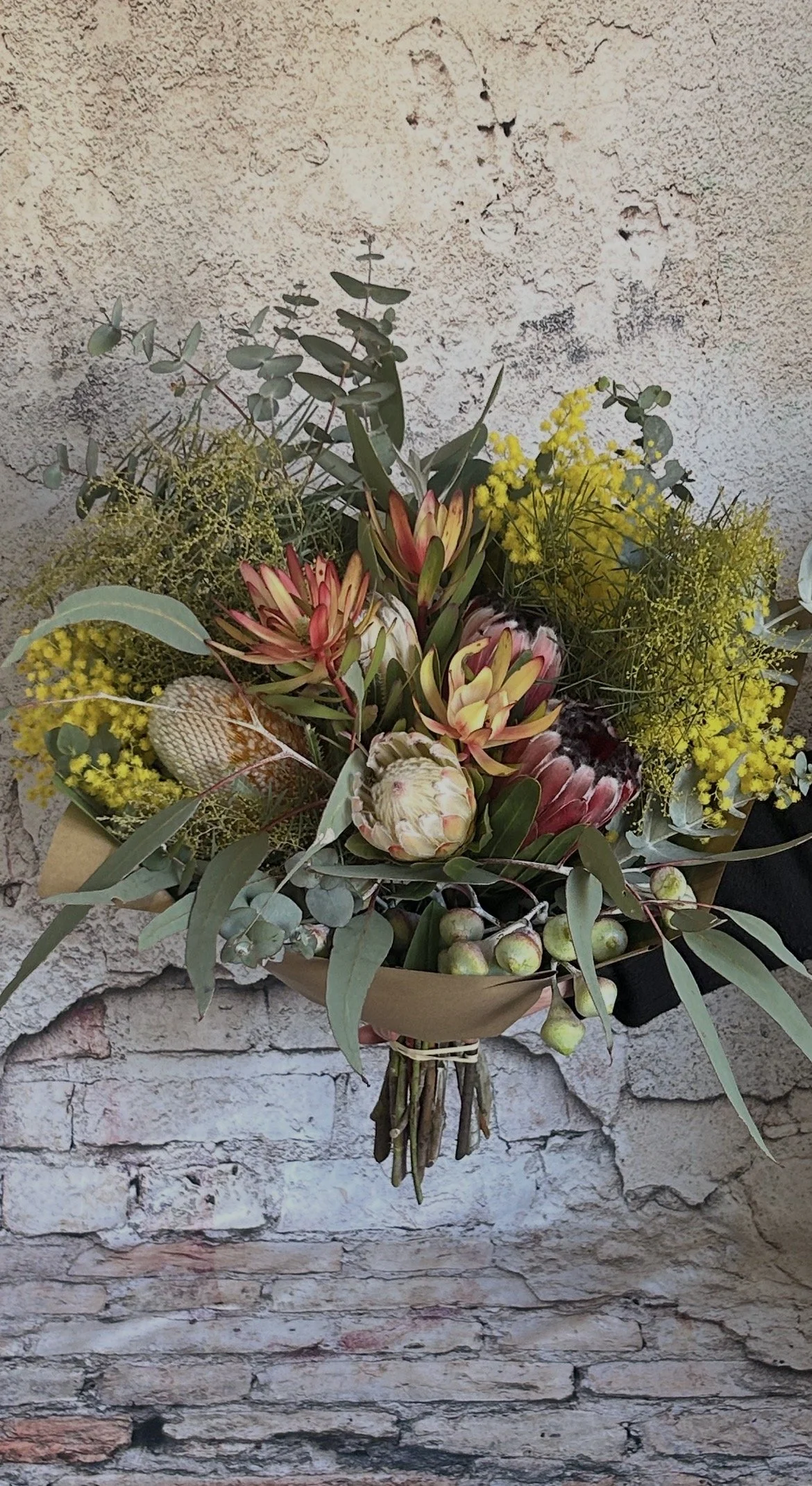 A bouquet of flowers with proteas, eucalyptus, and yellow blossoms held in front of a textured stone wall.
