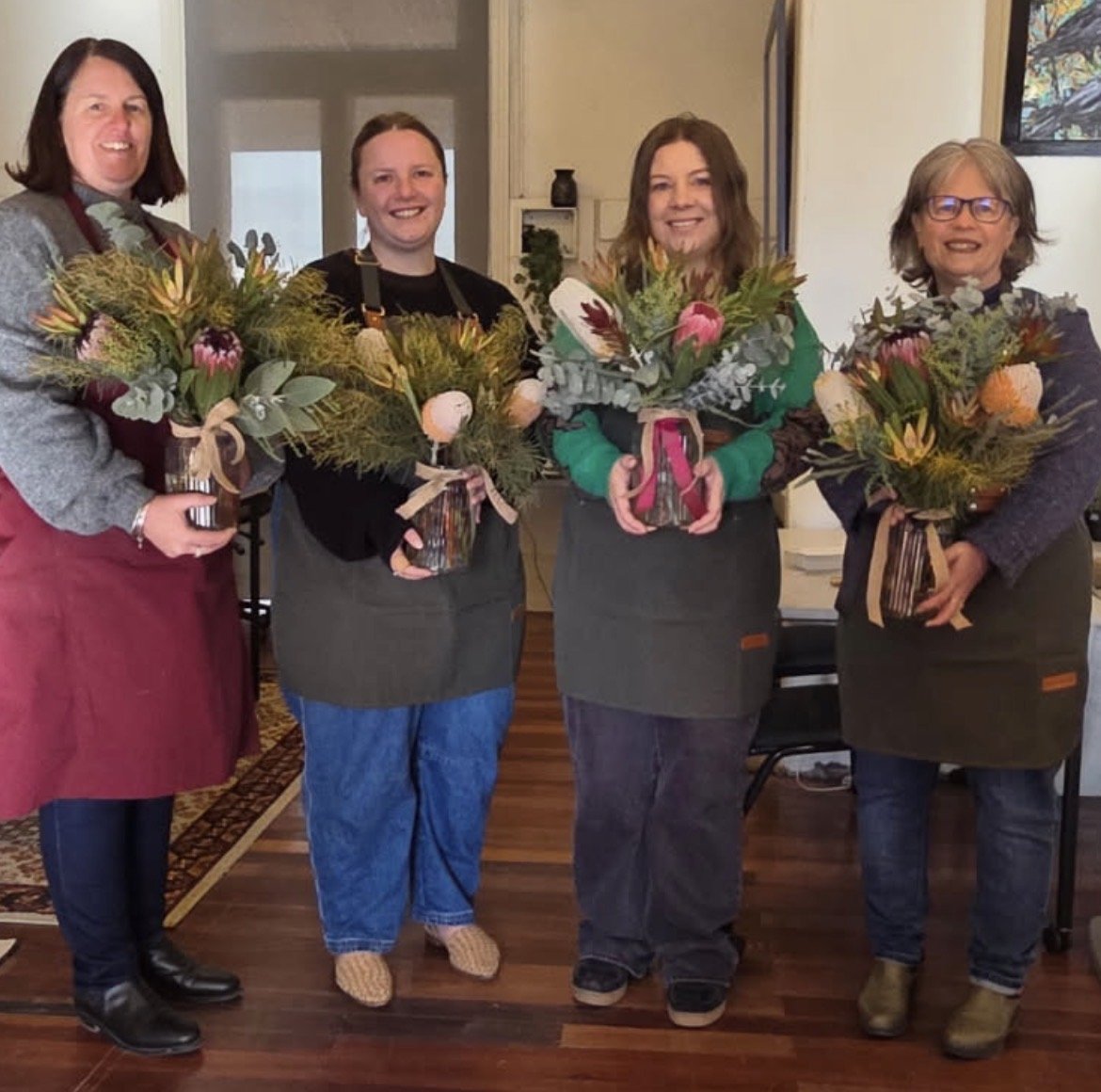 Four women standing indoors, each holding a floral arrangement in a decorated pot, smiling at the camera. They are wearing dark aprons, and the background shows a door, some shelves, and a framed picture.