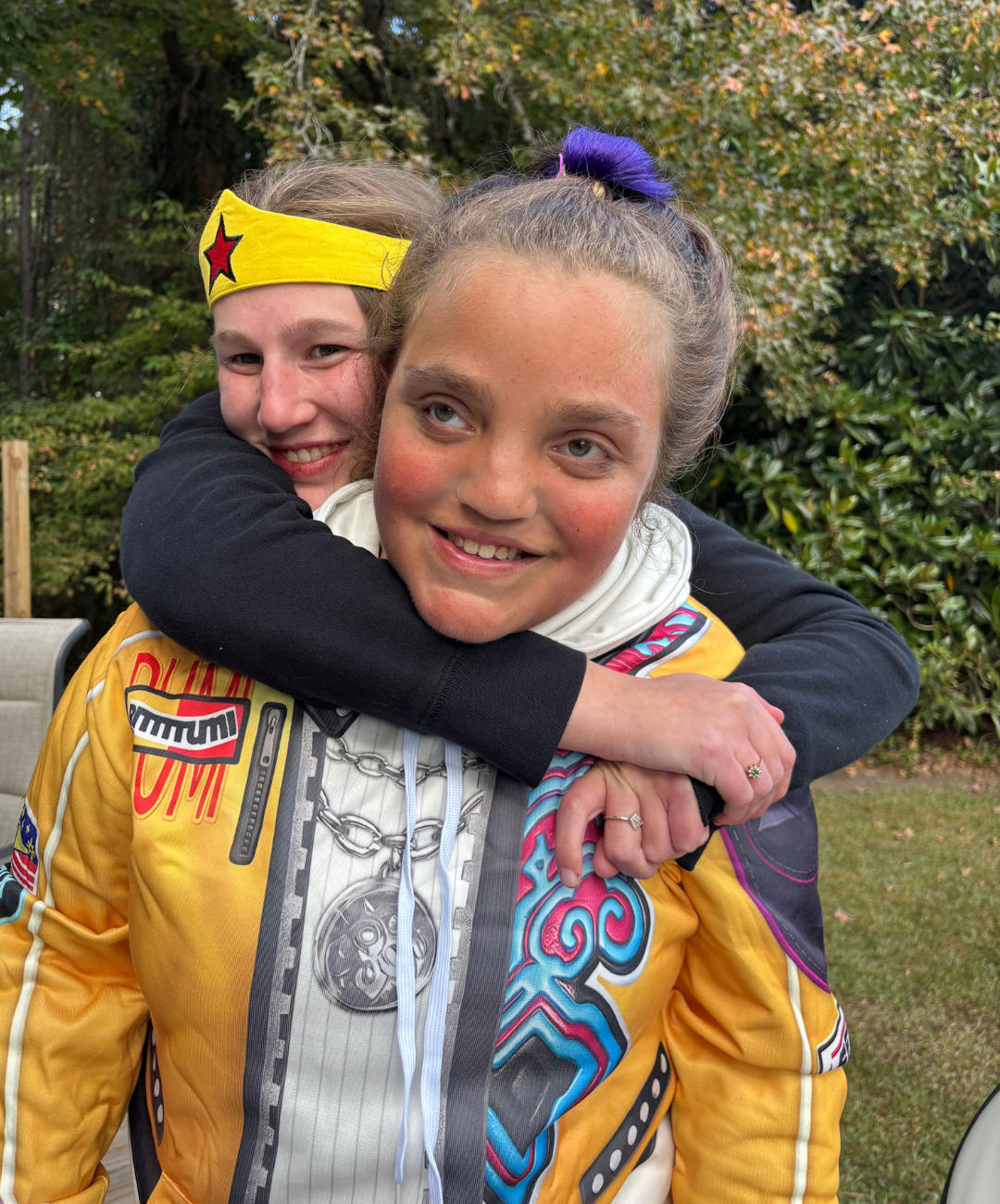Two smiling young girls hugging outdoors, one wearing a yellow racing jacket with patches and a yellow headband, the other with a purple-haired ponytail and black jacket, with greenery in the background.