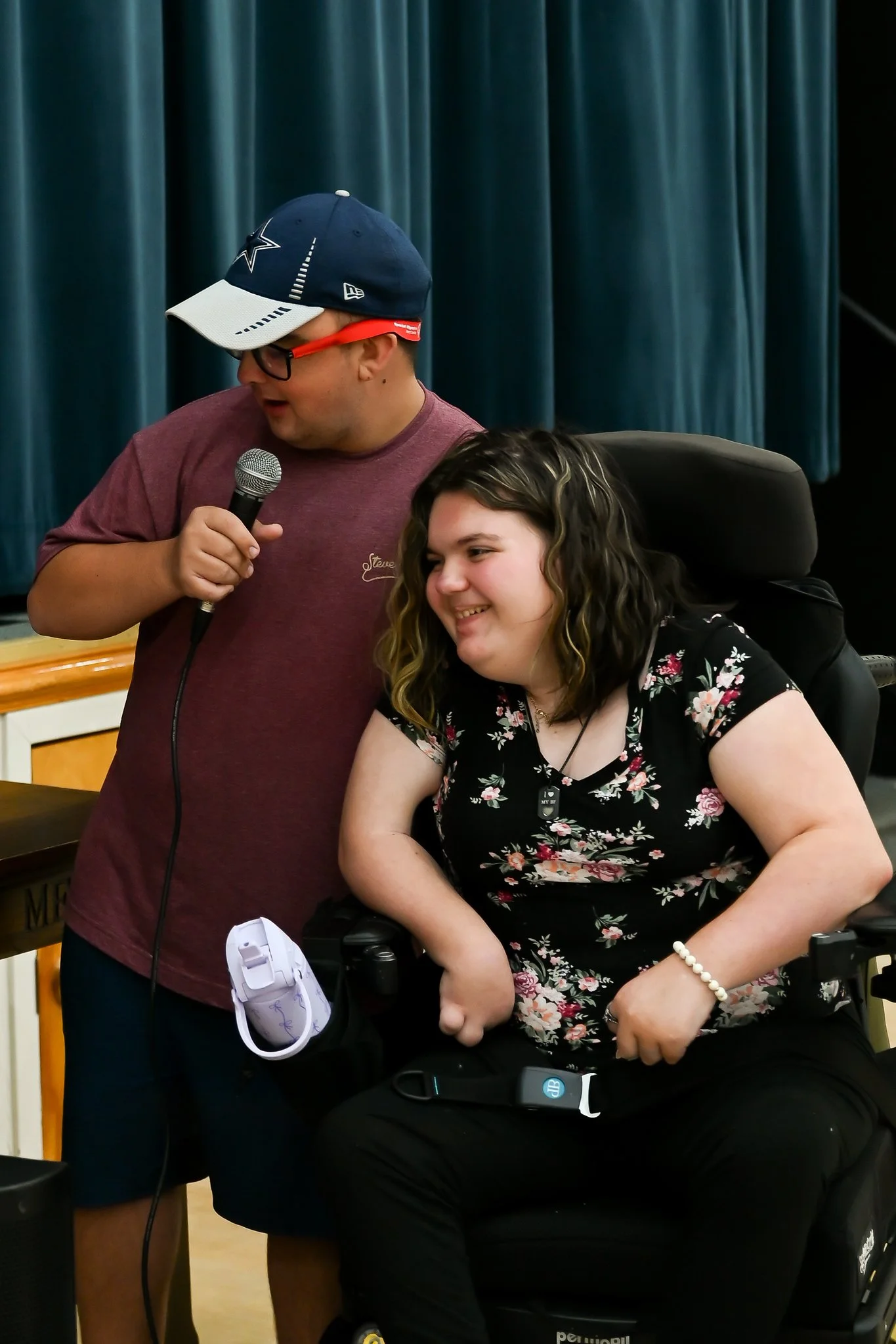 A man in a maroon shirt holding a microphone next to a woman in a wheelchair with curly hair, wearing a black floral dress, sitting in front of a blue curtain.