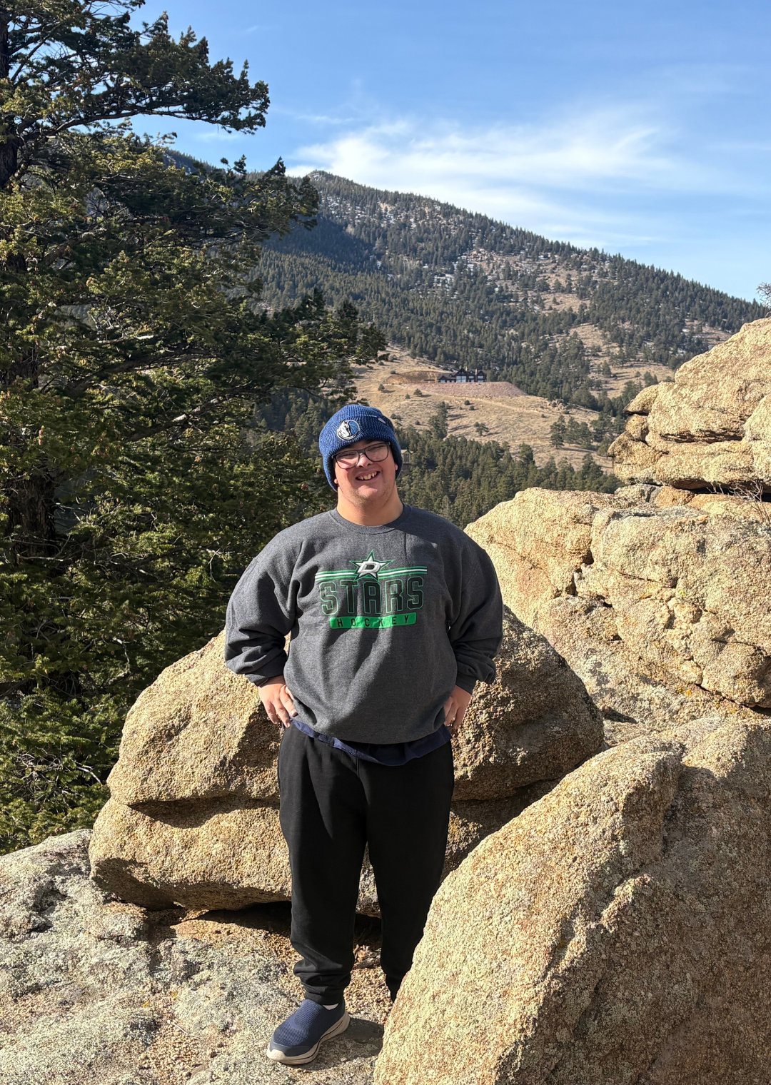 Man standing outdoors among large rocks with a mountainous forested background, wearing a blue beanie, glasses, a gray sweatshirt with "Stars Hockey" written on it, and black pants.