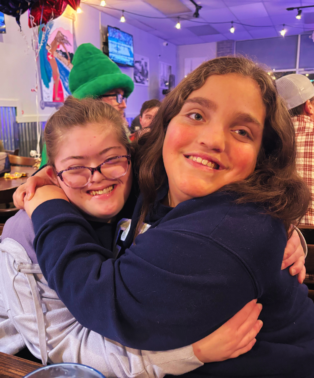 Two young women smiling and hugging at a lively indoor event, with colorful decorations and a TV screen in the background.