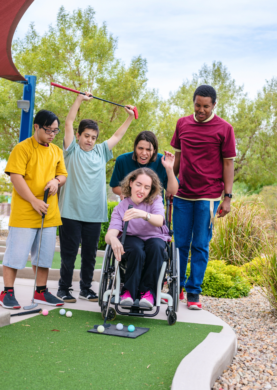Group of diverse people playing mini golf outdoors, with one woman in a wheelchair about to hit a golf ball, surrounded by others cheering and watching.
