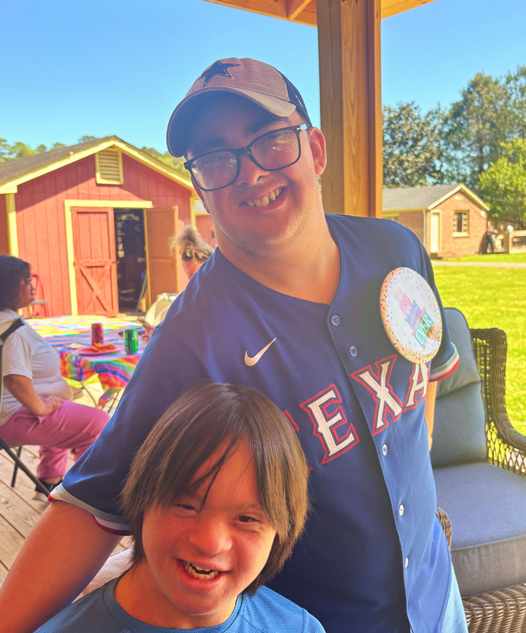 Two people smiling at an outdoor gathering on a porch, with a colorful table and a red shed in the background.