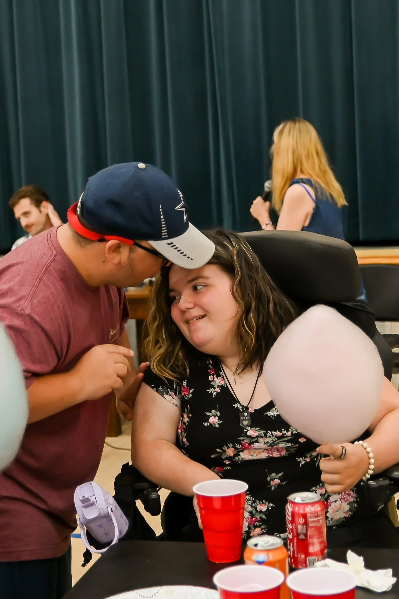 A woman with red hair and glasses is speaking into a microphone on a stage with dark green curtains. In the foreground, a man in a baseball cap and glasses is leaning in close to a young woman in a wheelchair, who is smiling and looking at him. There are red cups and cans of soda on the table in front of them, indicating a social gathering or celebration.