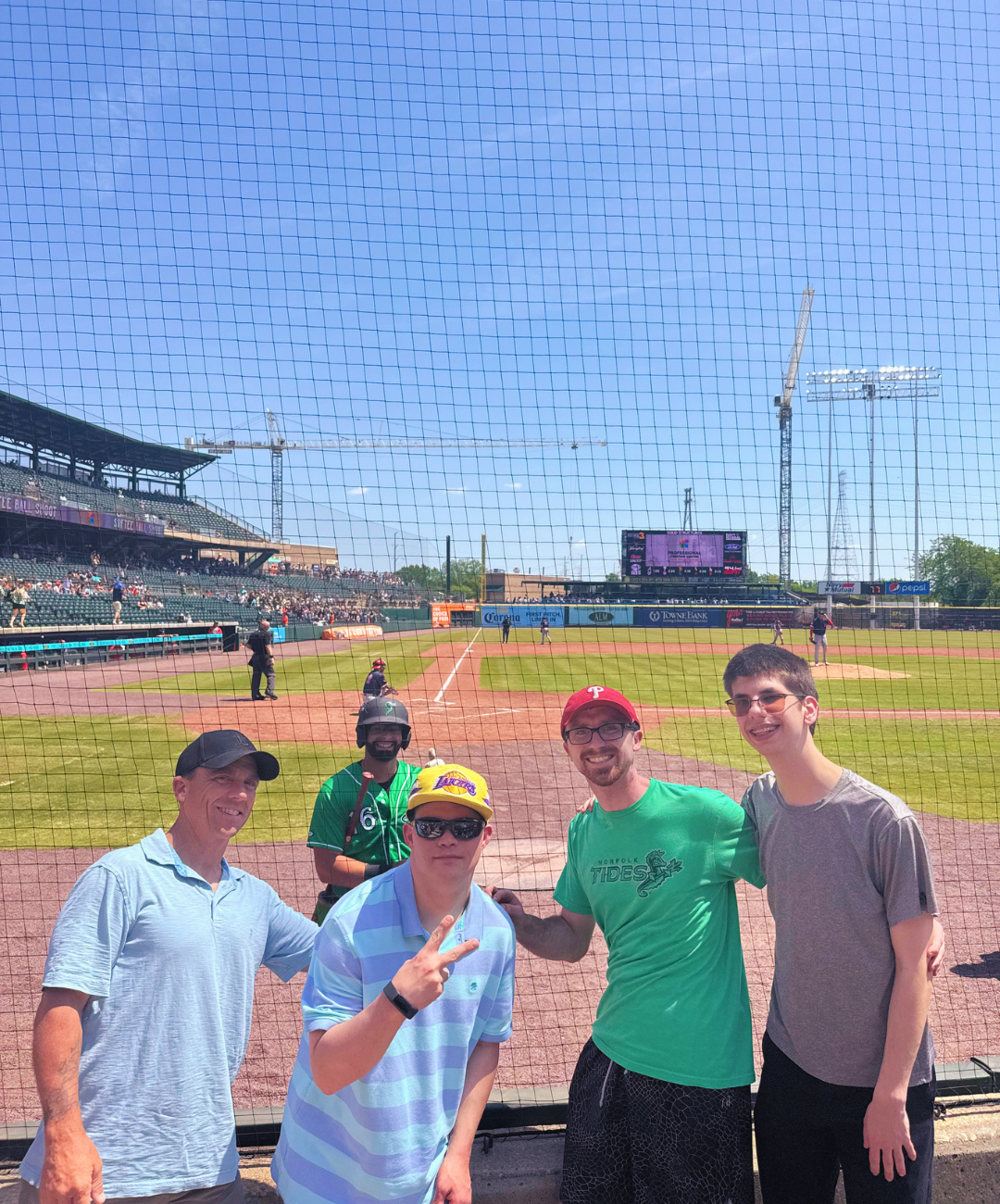Group of five men posing for a photo at a baseball stadium, with a baseball field, players, and a crowd in the background, during daytime.