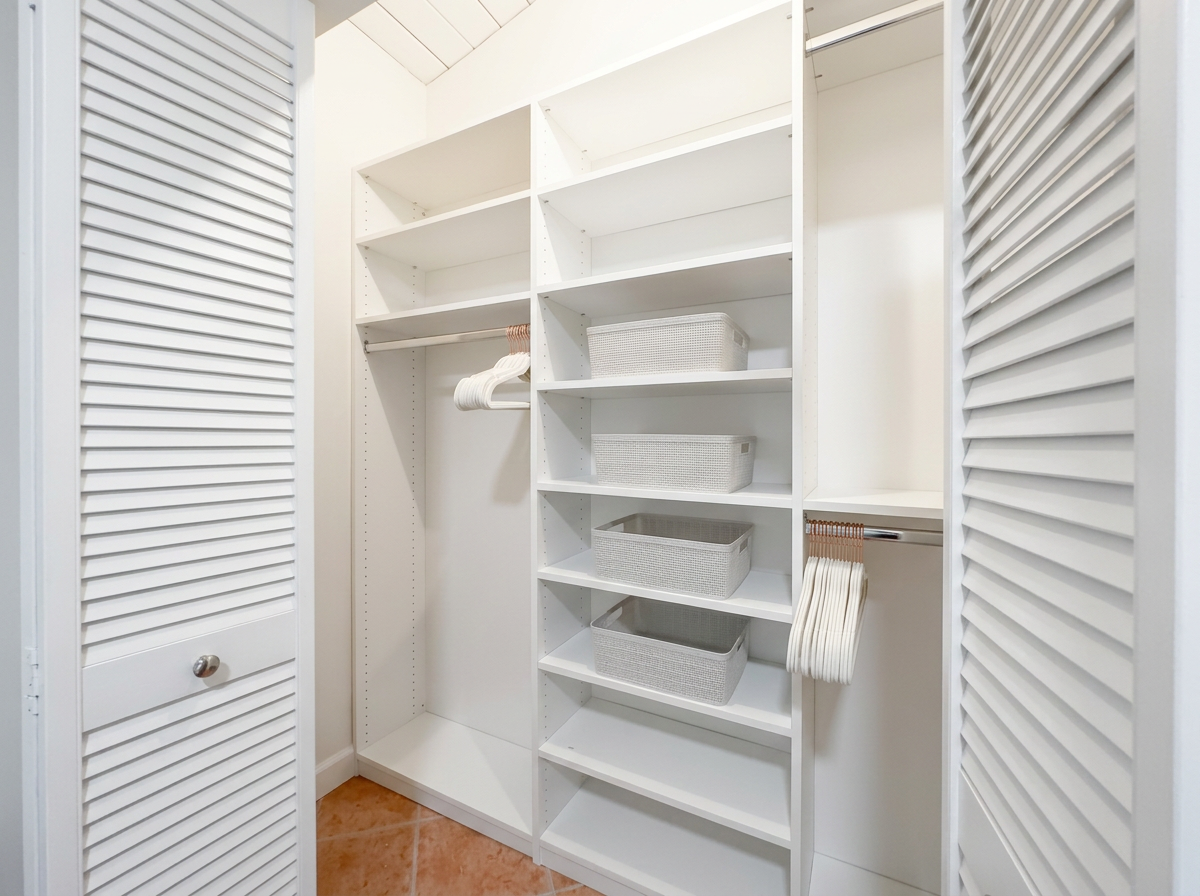 Empty closet with white shelving units, hanging rods, woven baskets, and napkin holders, with louvered closet doors on both sides.