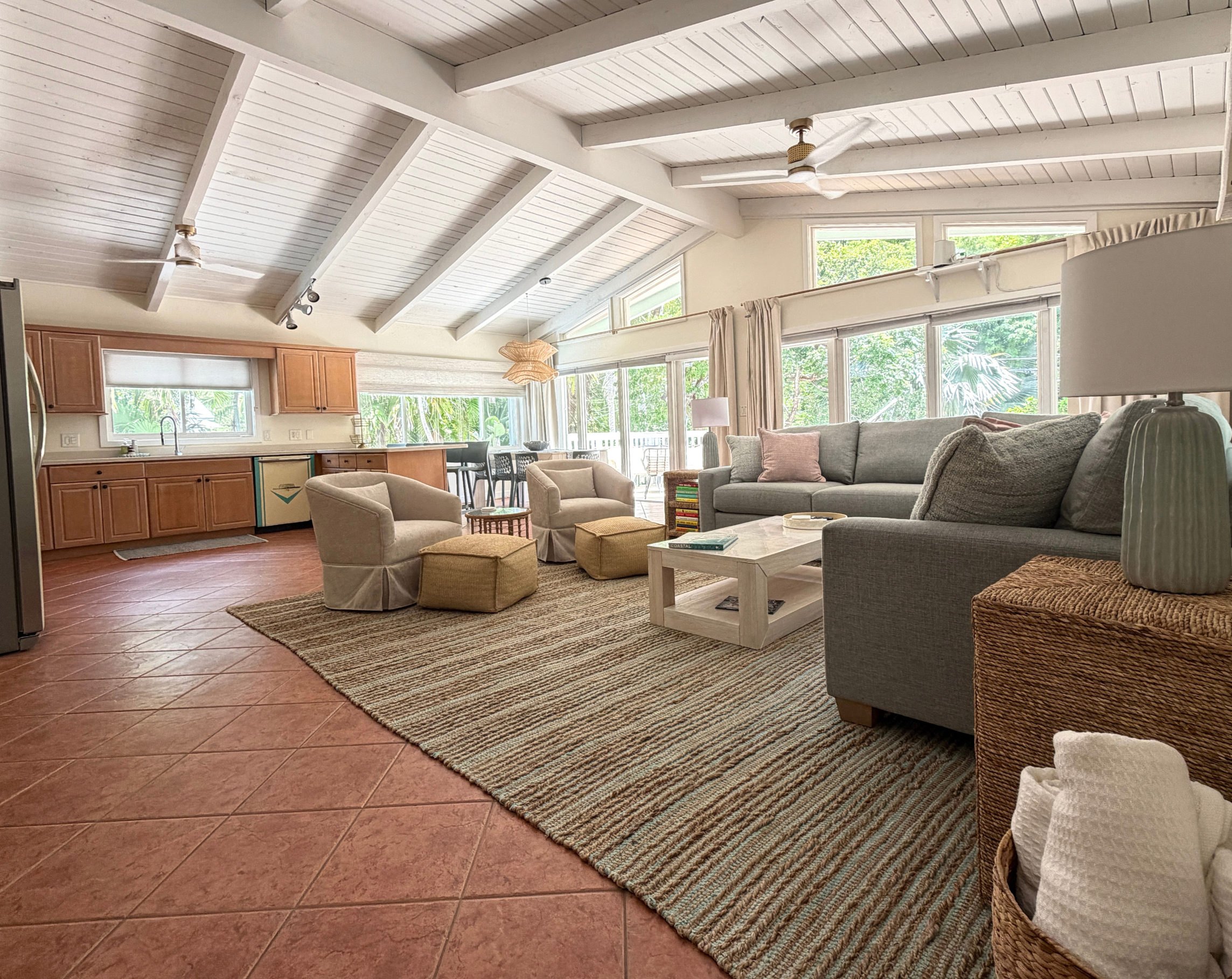 Living room with high vaulted ceiling, white wooden beams, large windows showing greenery, beige sofa with pink pillow, two beige armchairs, woven ottomans, wooden coffee table with books, kitchen area with wooden cabinets and stainless steel appliances, tiled floor, ceiling fans, and a rug.