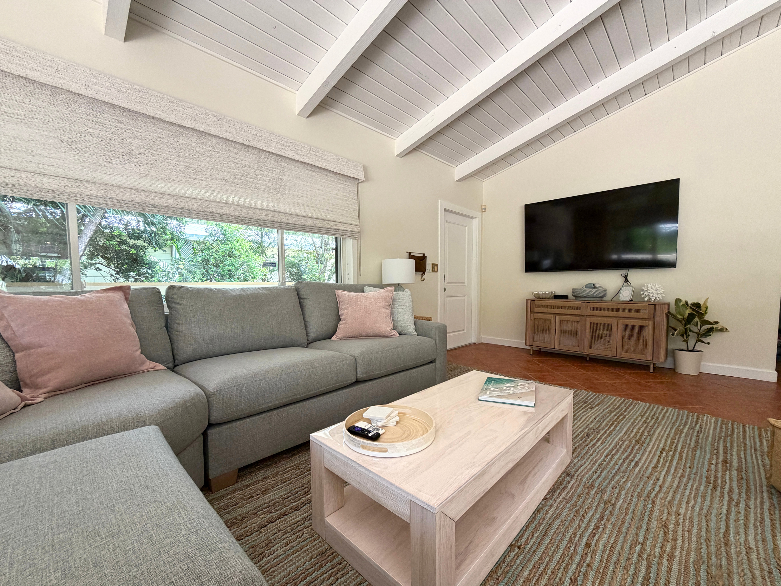Living room with a gray sofa with pink cushions, a wooden coffee table with a tray, TV mounted on wall, large window with blinds, and ceiling with white beams.