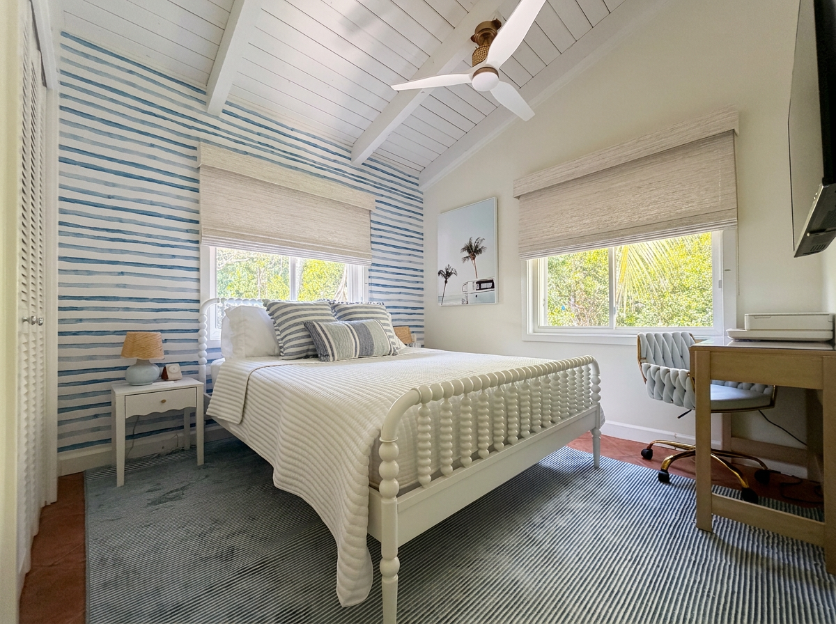 A bedroom with white walls and a blue and white striped accent wall, a white bed with striped pillows, two windows with beige roman shades, a small white bedside table with a lamp, a desk with a gray tufted office chair, and a ceiling fan.
