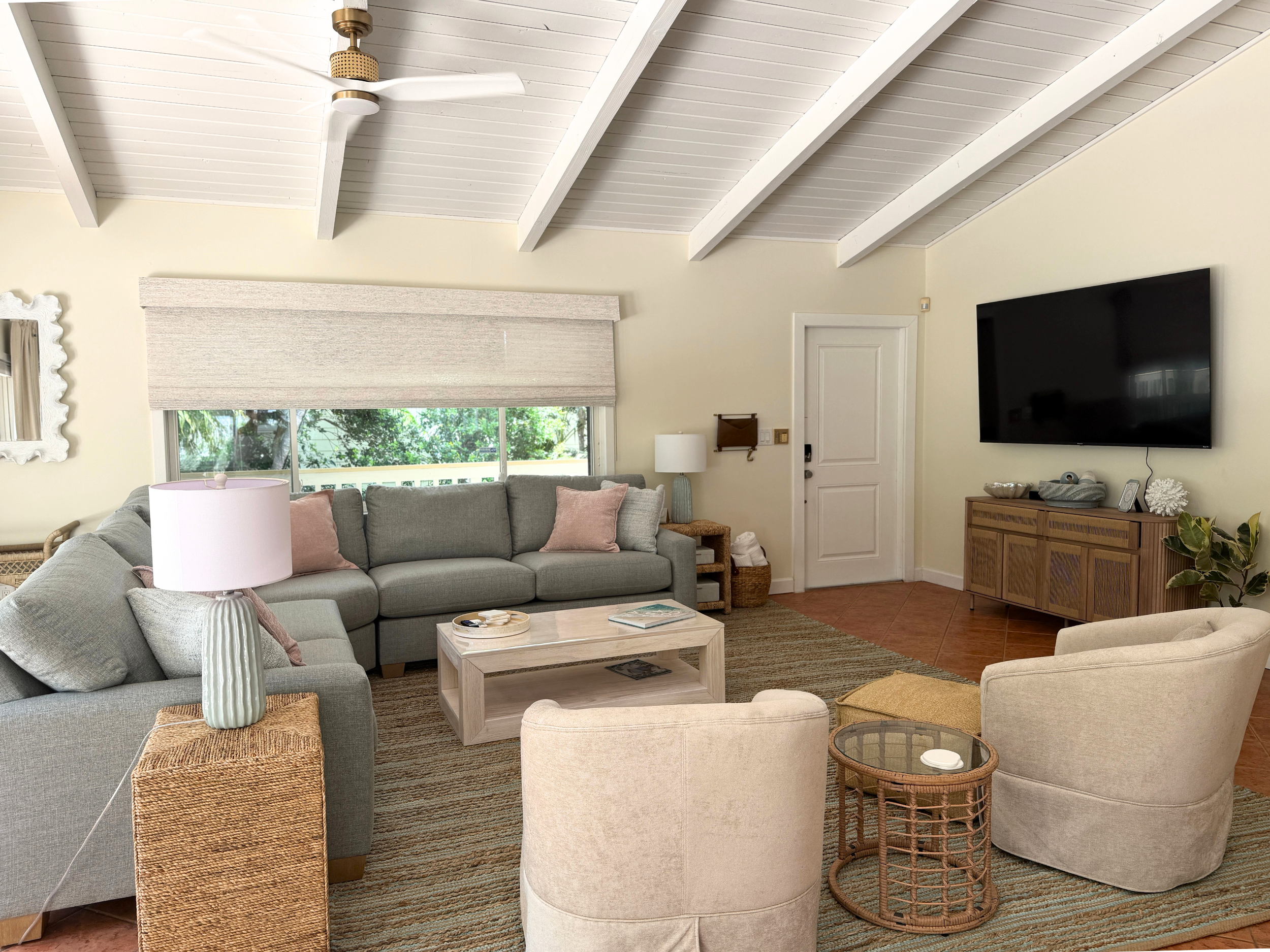Living room with beige walls and white vaulted ceiling, large window with blinds, gray sectional sofa with pink pillows, framed mirror, white tabletop lamp, wooden side table, large flat-screen TV, wicker and fabric ottomans, small plant, and wooden cabinet.
