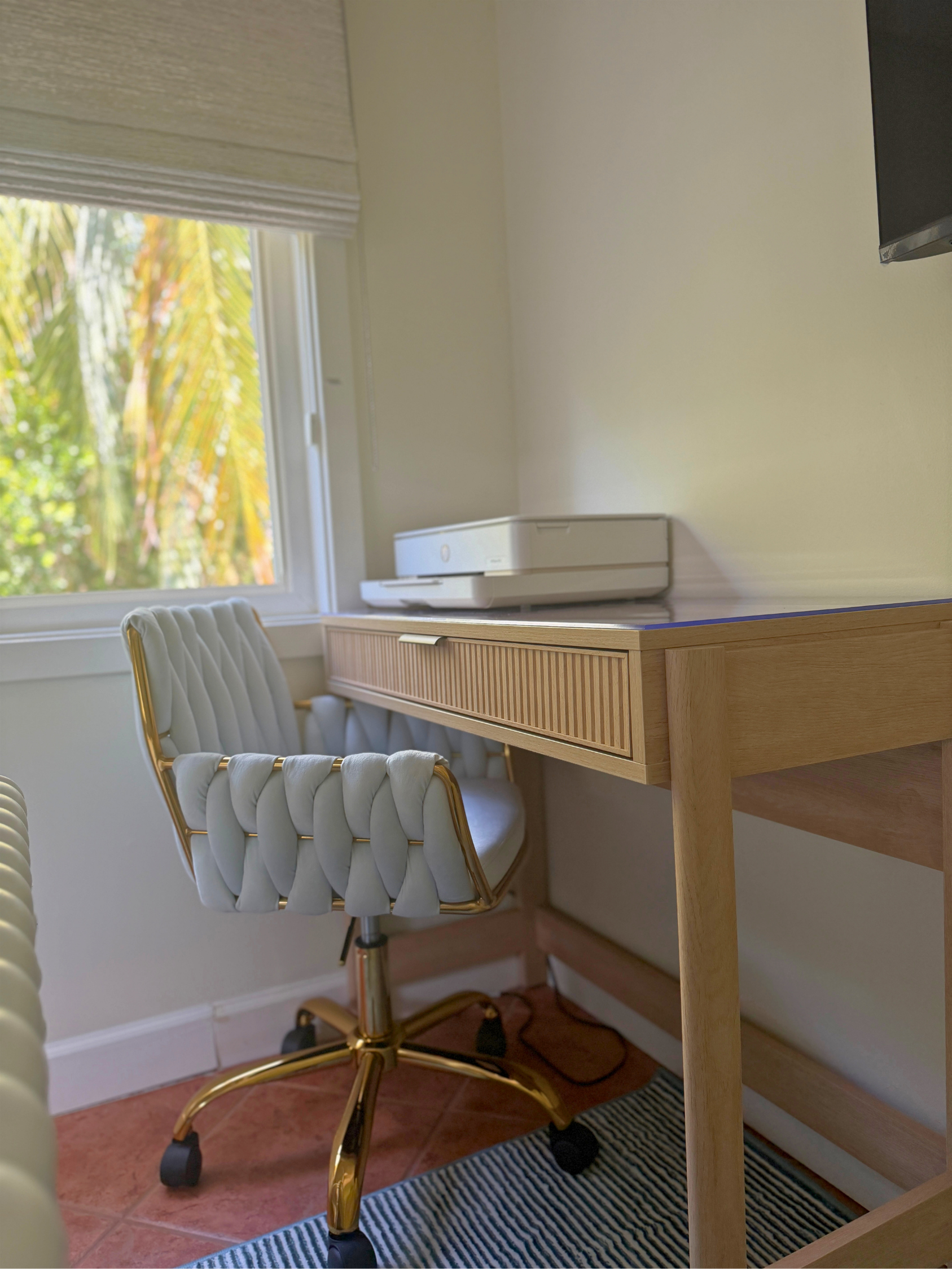 A white office chair with a gold frame and white padding, positioned next to a light wood desk in a room with a window showing green foliage outside. There is a printer on the desk, and the floor is covered with a striped black and white rug.