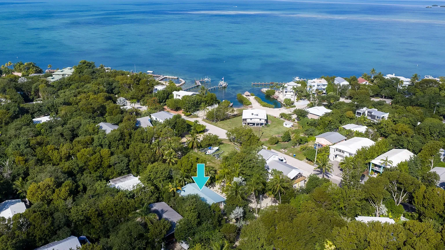 Aerial view of a coastal residential area with many houses, trees, and a water body with docks and boats in the background.