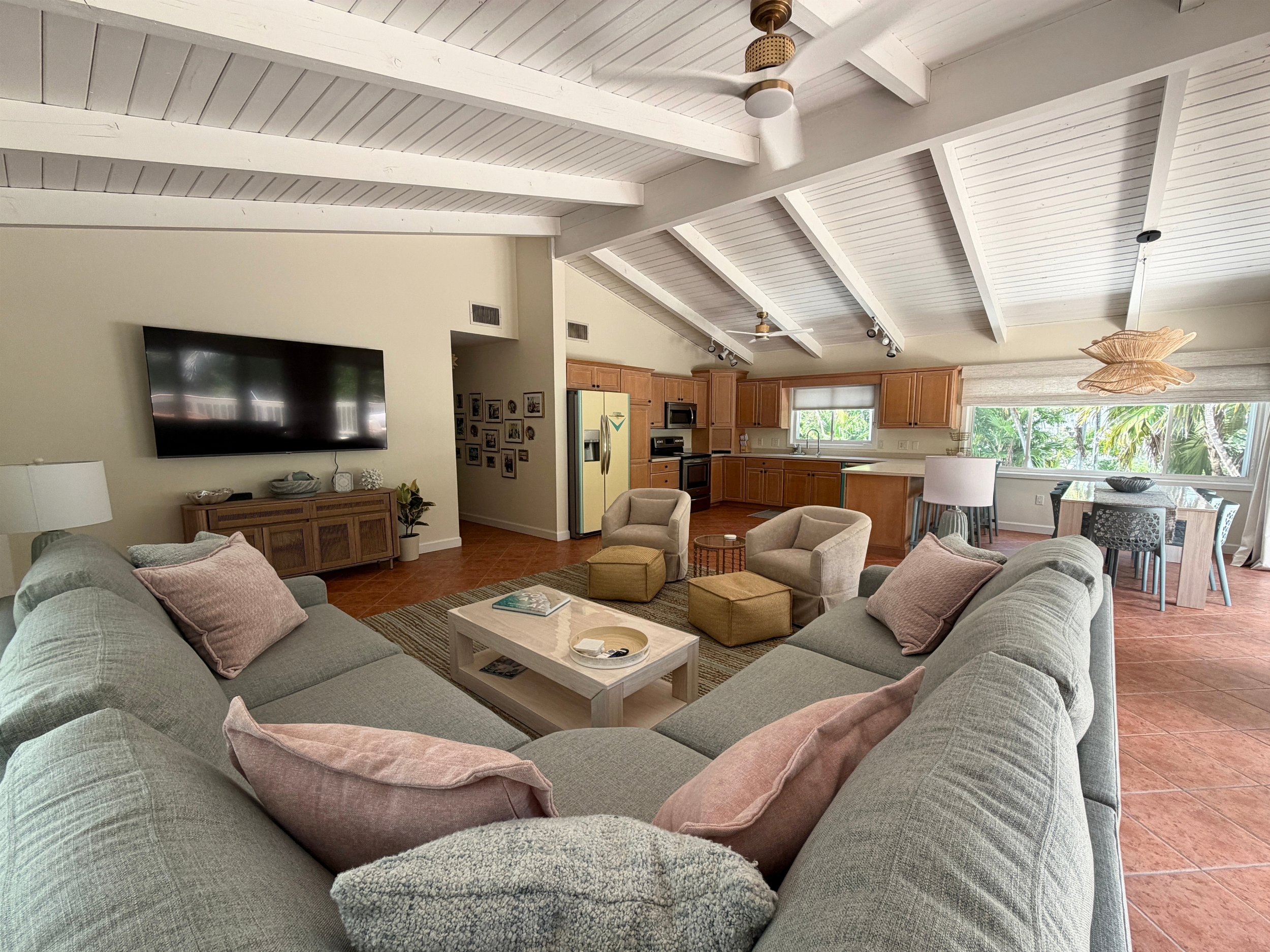 Living room with a large sectional sofa, a coffee table, and decorative pillows, connected to a kitchen area with wooden cabinets and a window showing greenery outside.