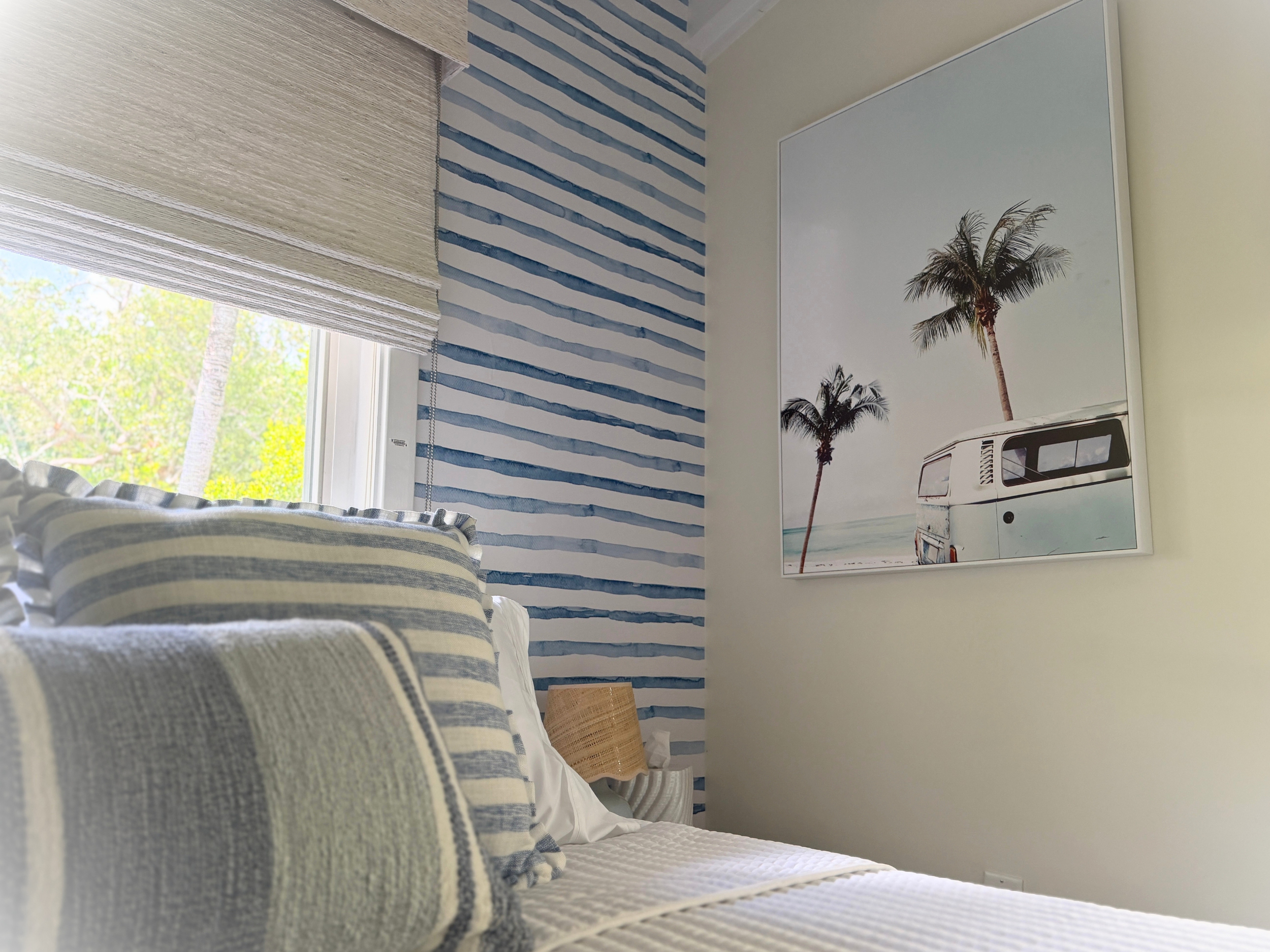 Interior view of a bedroom with a window, curtains, striped blue and white wallpaper, a painting of palm trees, and a bed with a striped blanket.