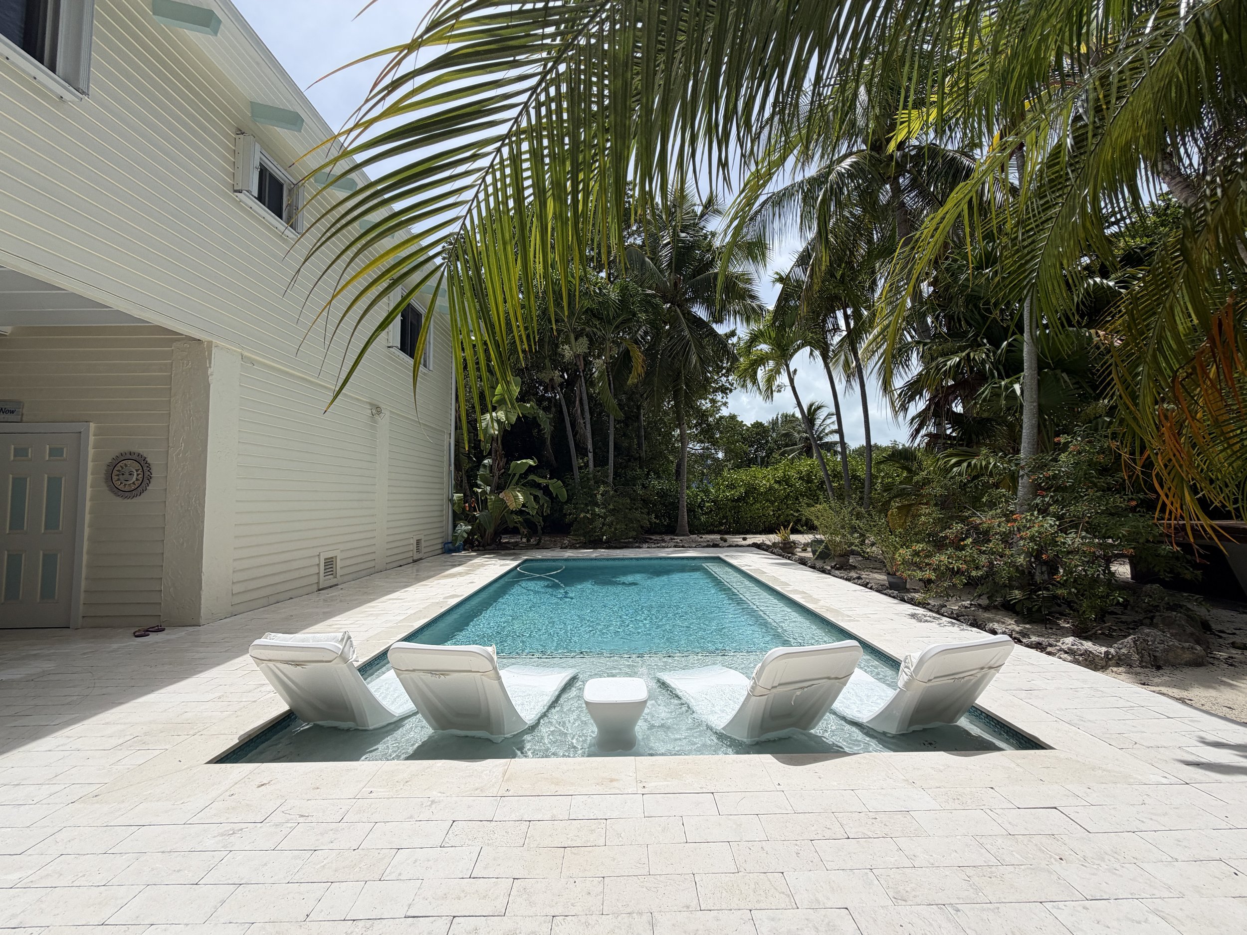 Swimming pool surrounded by white tile deck, with four white lounge chairs, a white building with sliding doors, and lush green palm trees in the Florida Keys.