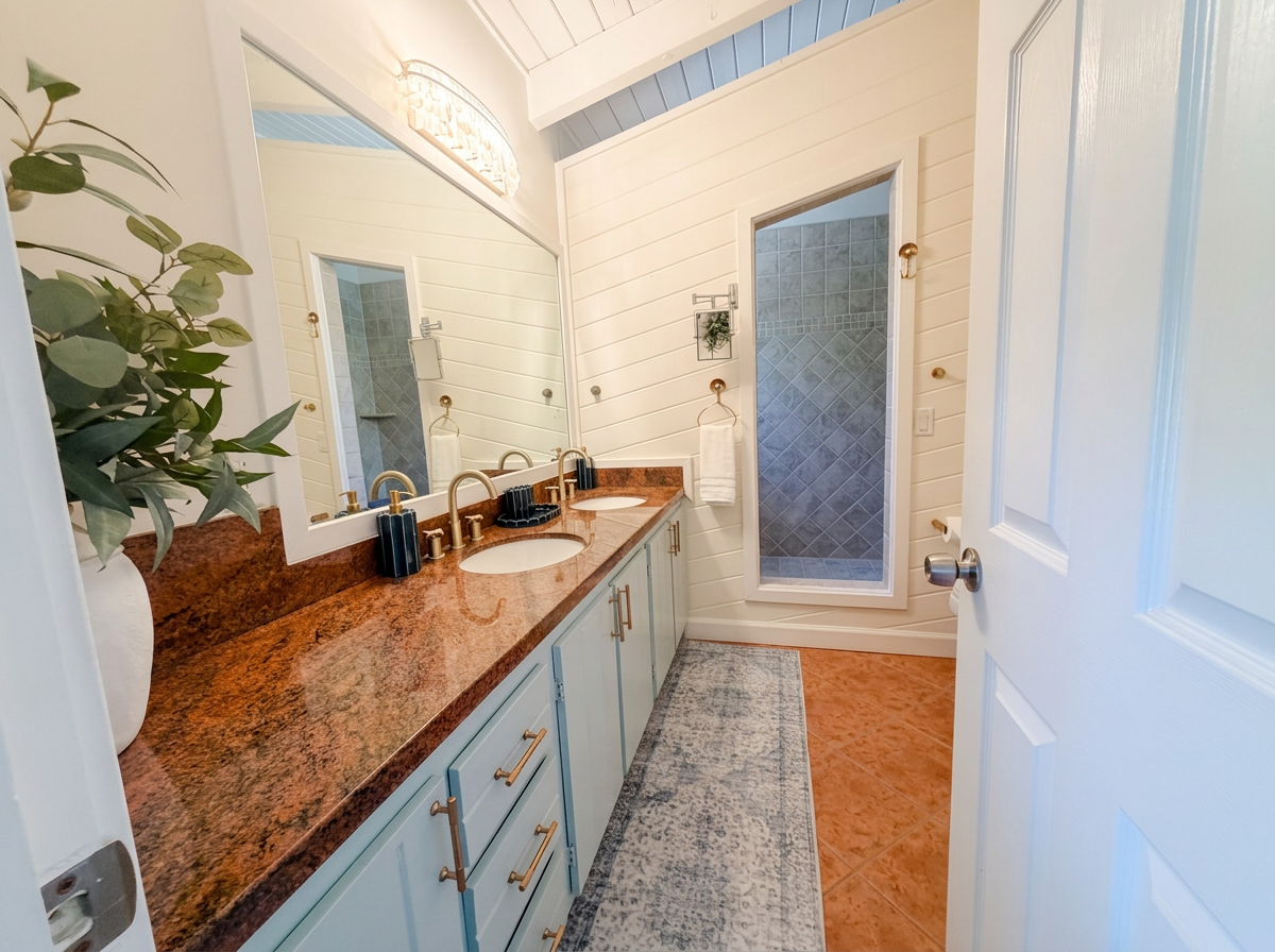 Bathroom with double sink vanity featuring a granite countertop, a large mirror, and black accents. There is a potted plant near the door, a textured rug on the floor, and a walk-in shower with tile walls visible through an open doorway.
