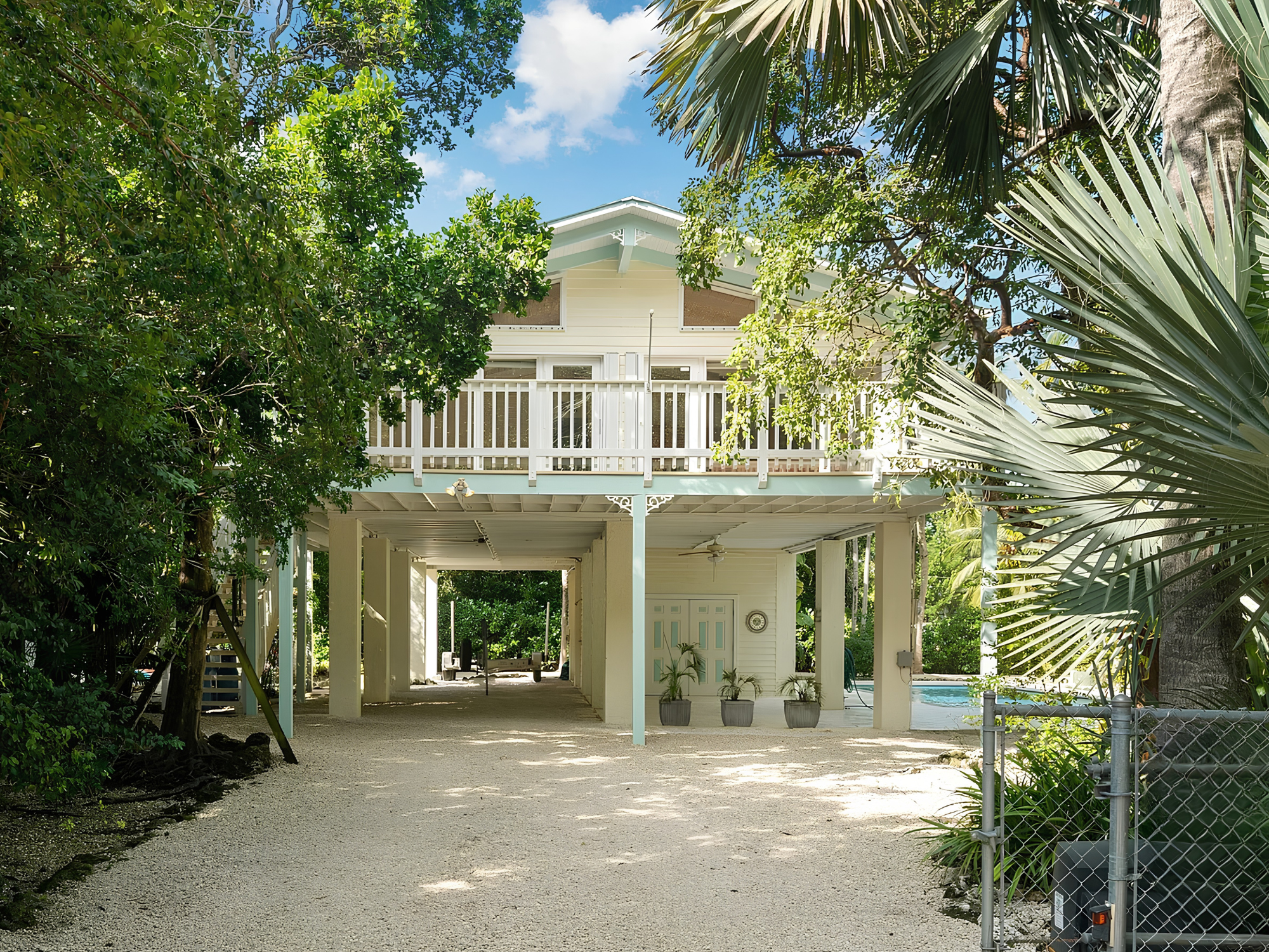 View of a house with a large porch surrounded by lush green trees and plants, with a bright blue sky and clouds visible.
