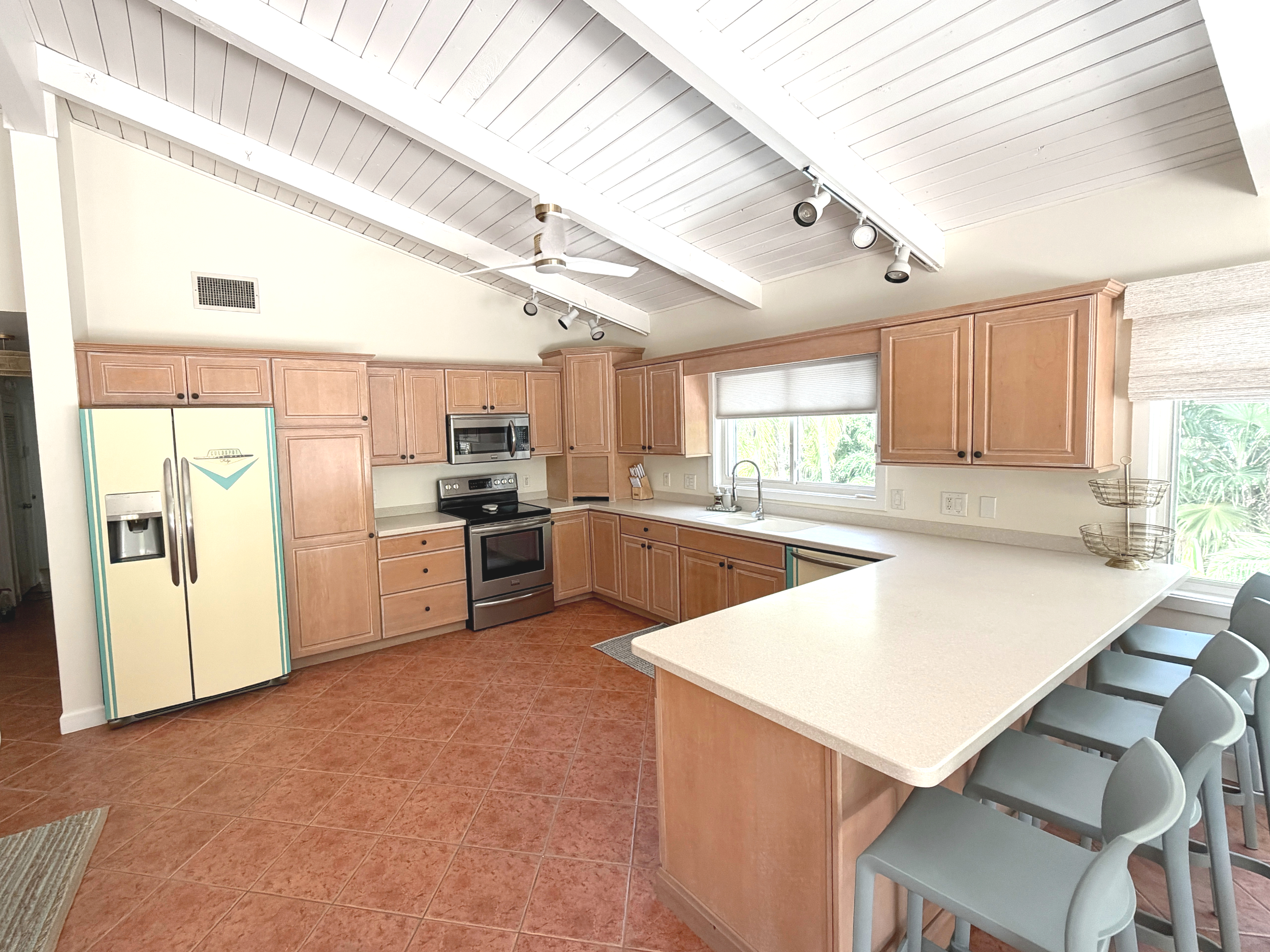 A kitchen with a vaulted white beadboard ceiling, wooden cabinets, stainless steel appliances, and a breakfast bar with gray chairs. There are large windows allowing natural light in.