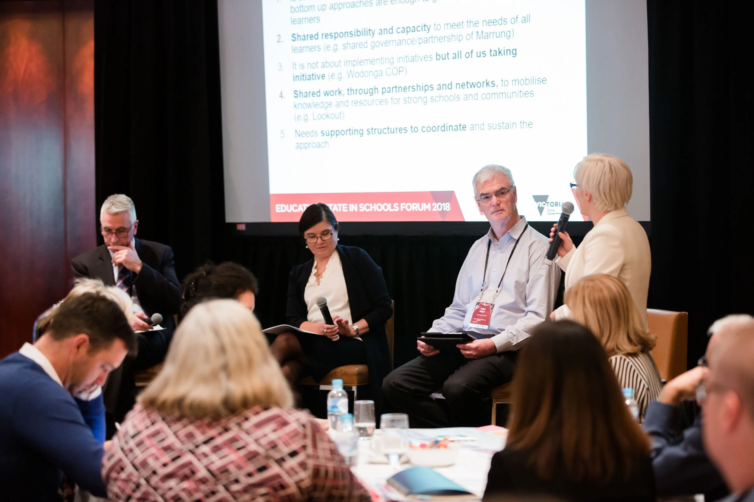 A group of people participating in a panel discussion during a conference or forum, with a large projected presentation slide titled 'Educate State in Schools Forum 2018' in the background. The panel includes a woman holding a microphone and three men, with an audience seated in front of them.