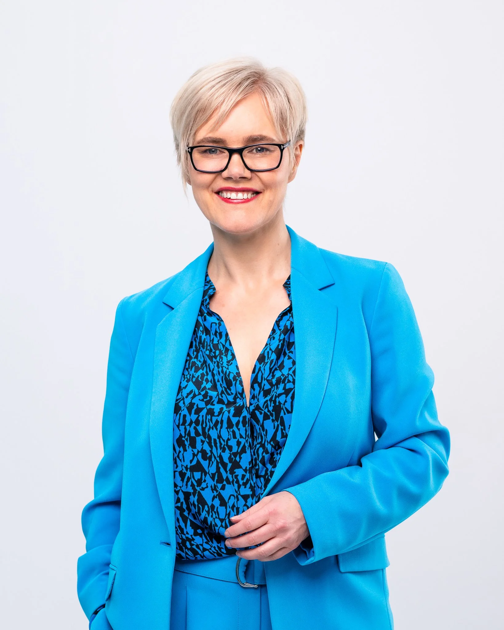 A woman with short blonde hair, glasses, and a bright smile, wearing a blue blazer over a patterned blue and black blouse, standing against a plain white background.