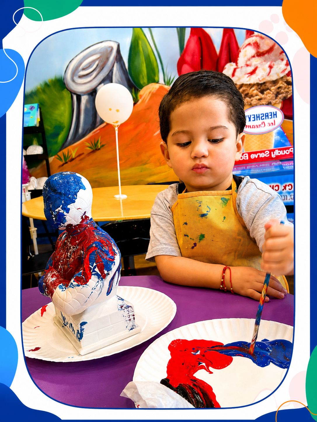 A young boy wearing a yellow apron is painting a plaster bust with red, blue, and white paint at a party or craft station.