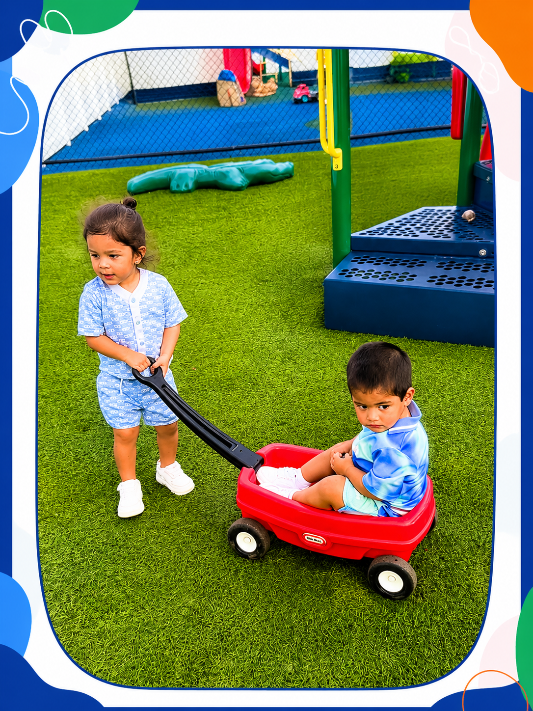 Two young children playing outdoors on artificial grass, one girl standing and pulling a red wagon with a boy sitting inside, at a playground with play structures in the background.
