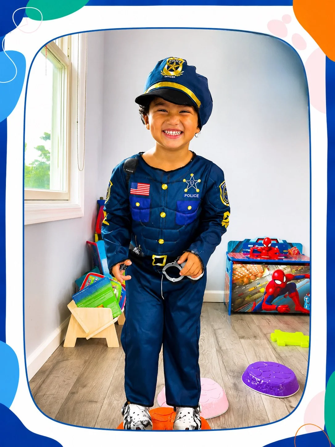 Young boy dressed as a police officer, smiling, standing indoors with toys and a Spider-Man-themed storage box in the background.