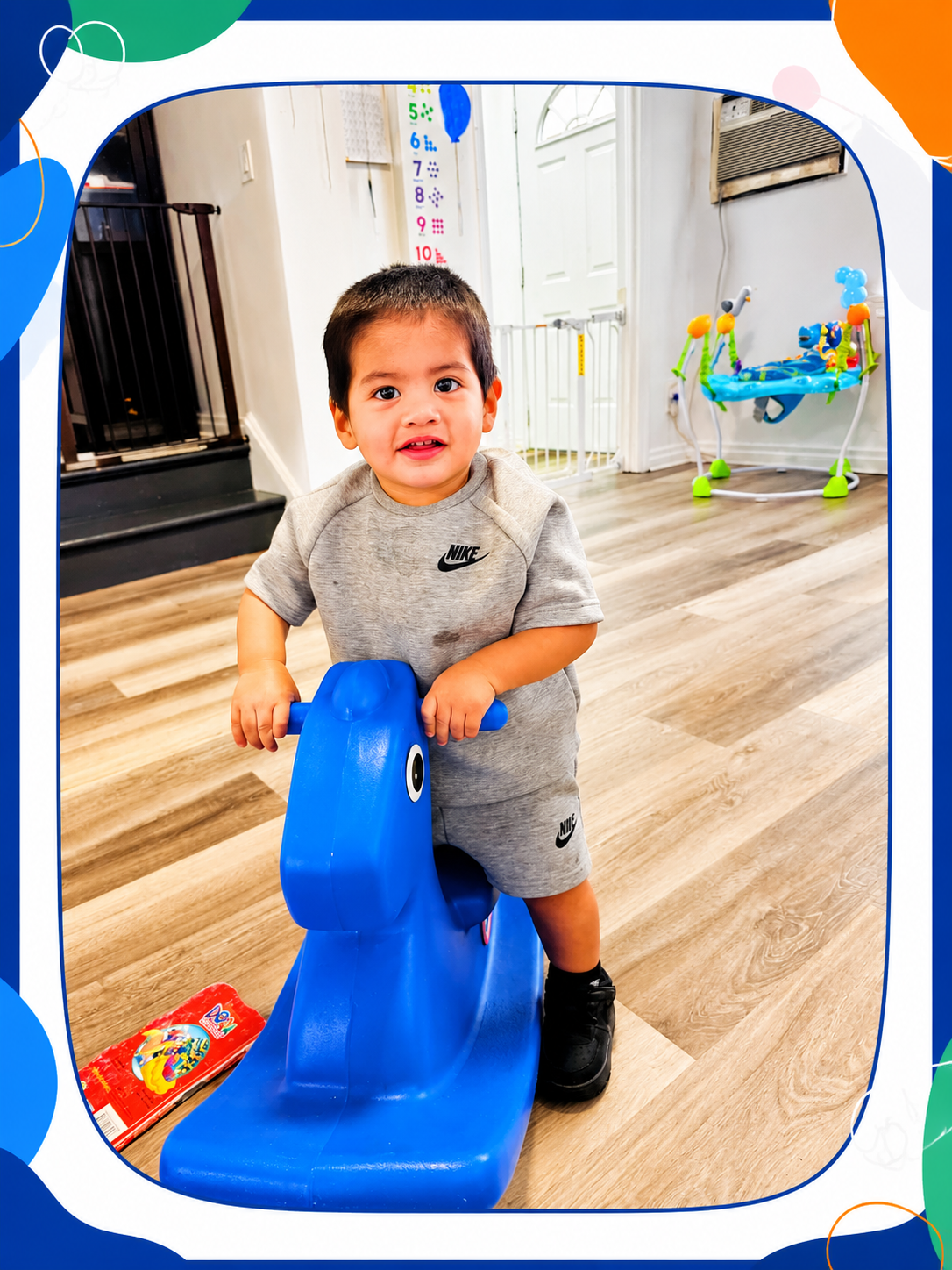 Young boy riding a blue rocking horse in a playroom, with a colorful toy and height chart in the background.