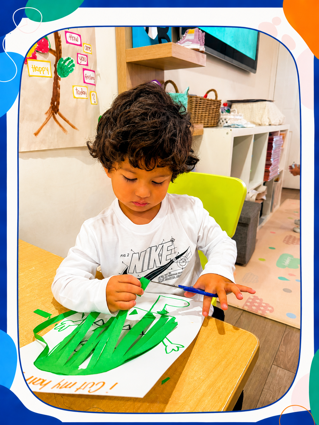 A young boy with curly hair coloring a paper craft project with green paper strips and a blue marker at a classroom table.