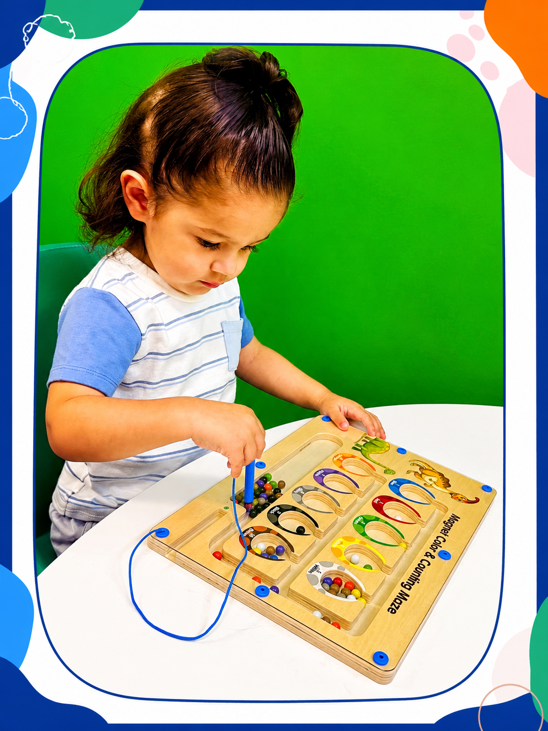 Young girl playing with a marble maze toy with colorful paths and marbles at a white table against a green background.