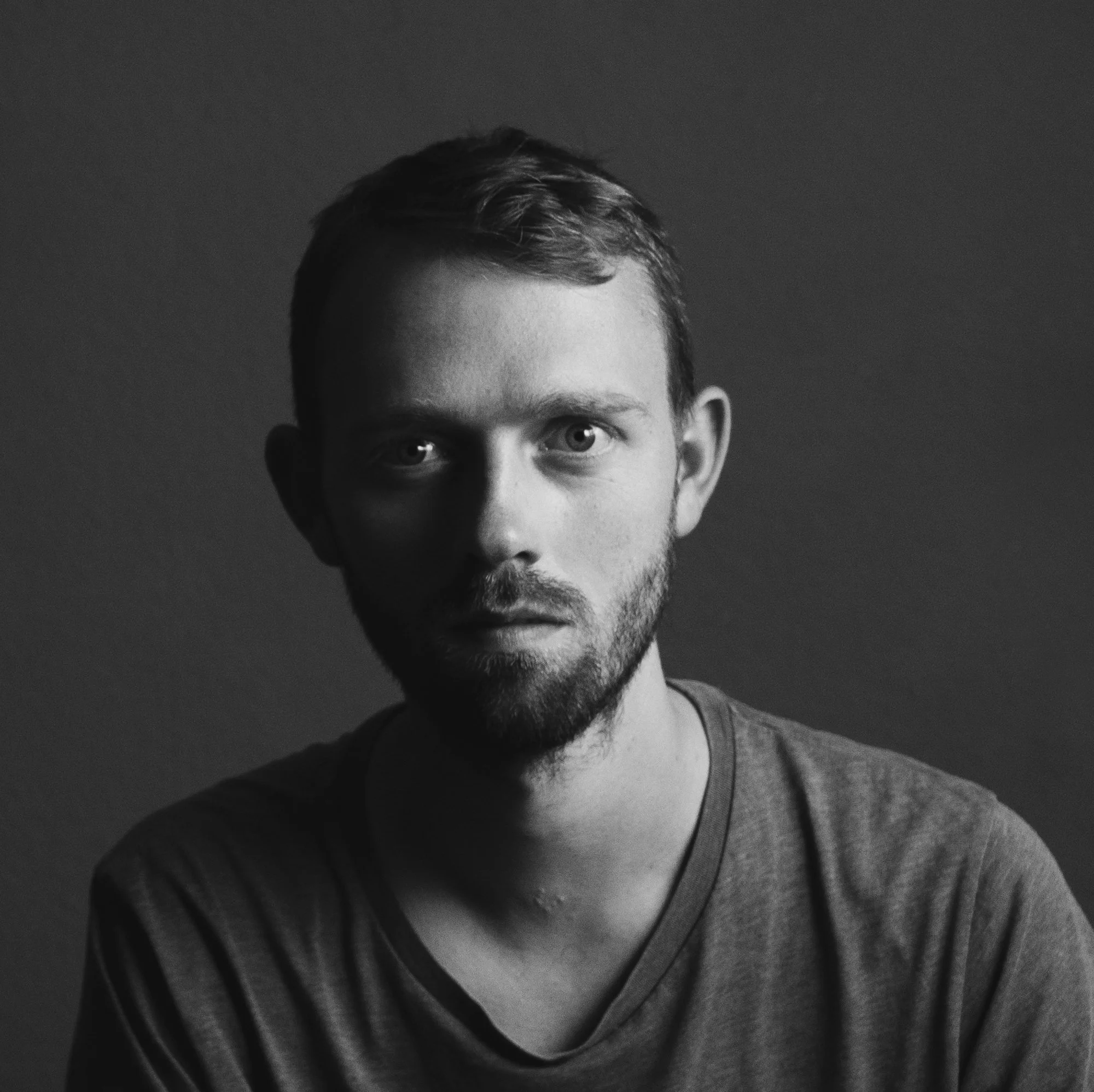 Black and white portrait of a young man with short hair, beard, wearing a dark shirt, with a shadowed background.