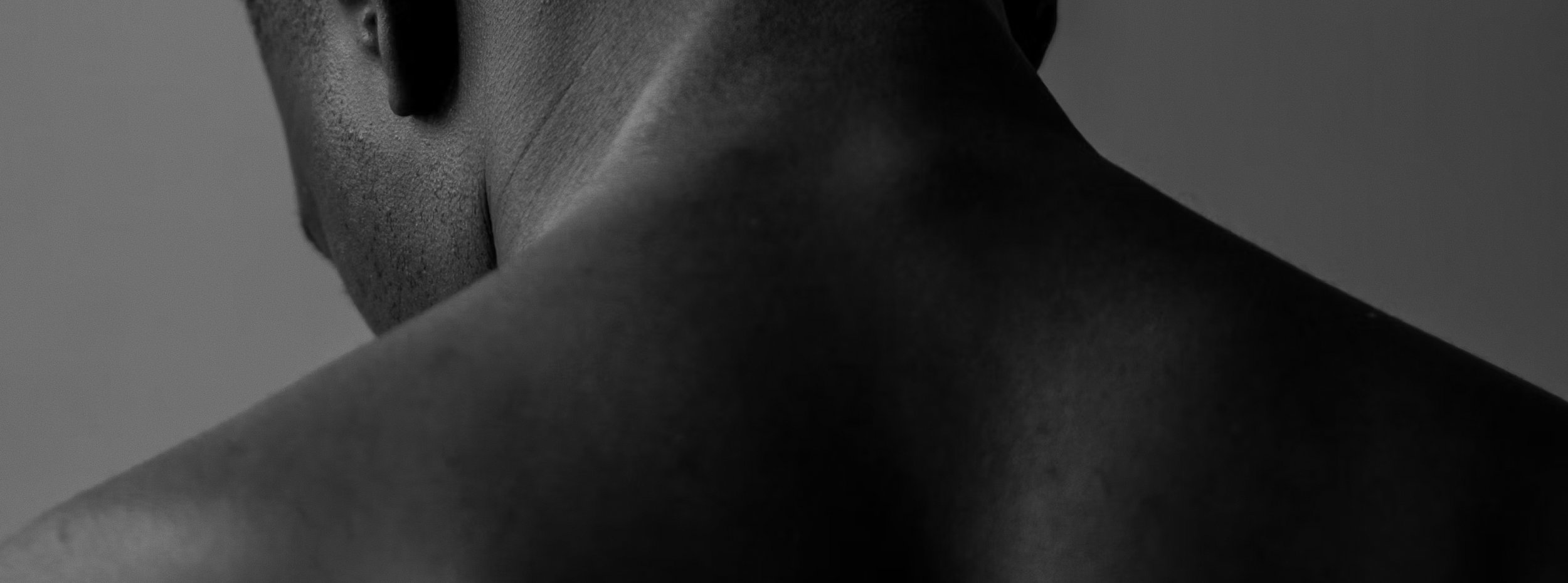 Close-up black and white photograph of a person's neck and shoulder from behind, showing textured skin and short hair.